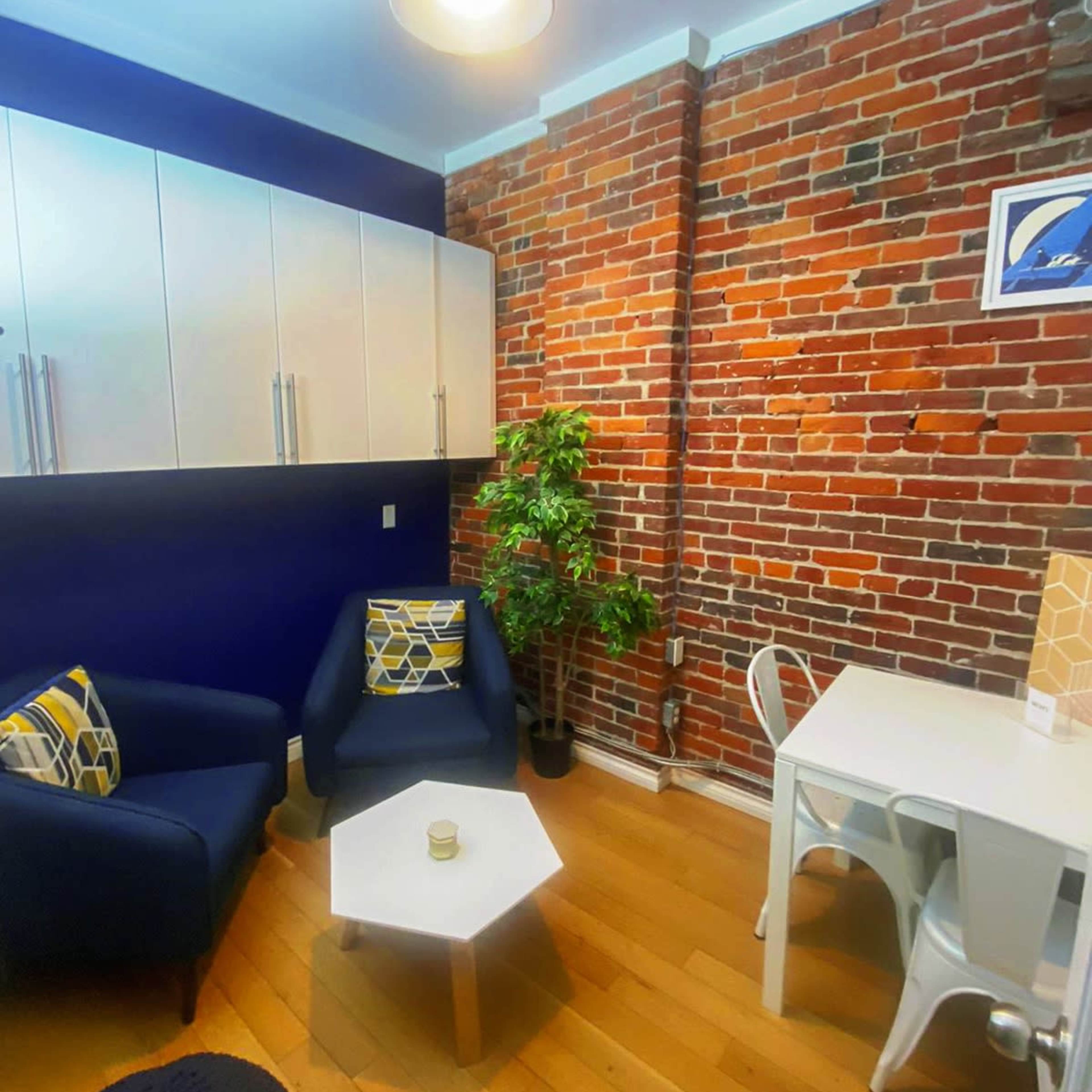 A small living space featuring a brick wall, a blue sofa, a small coffee table, a white dining table with chairs, and a potted plant.