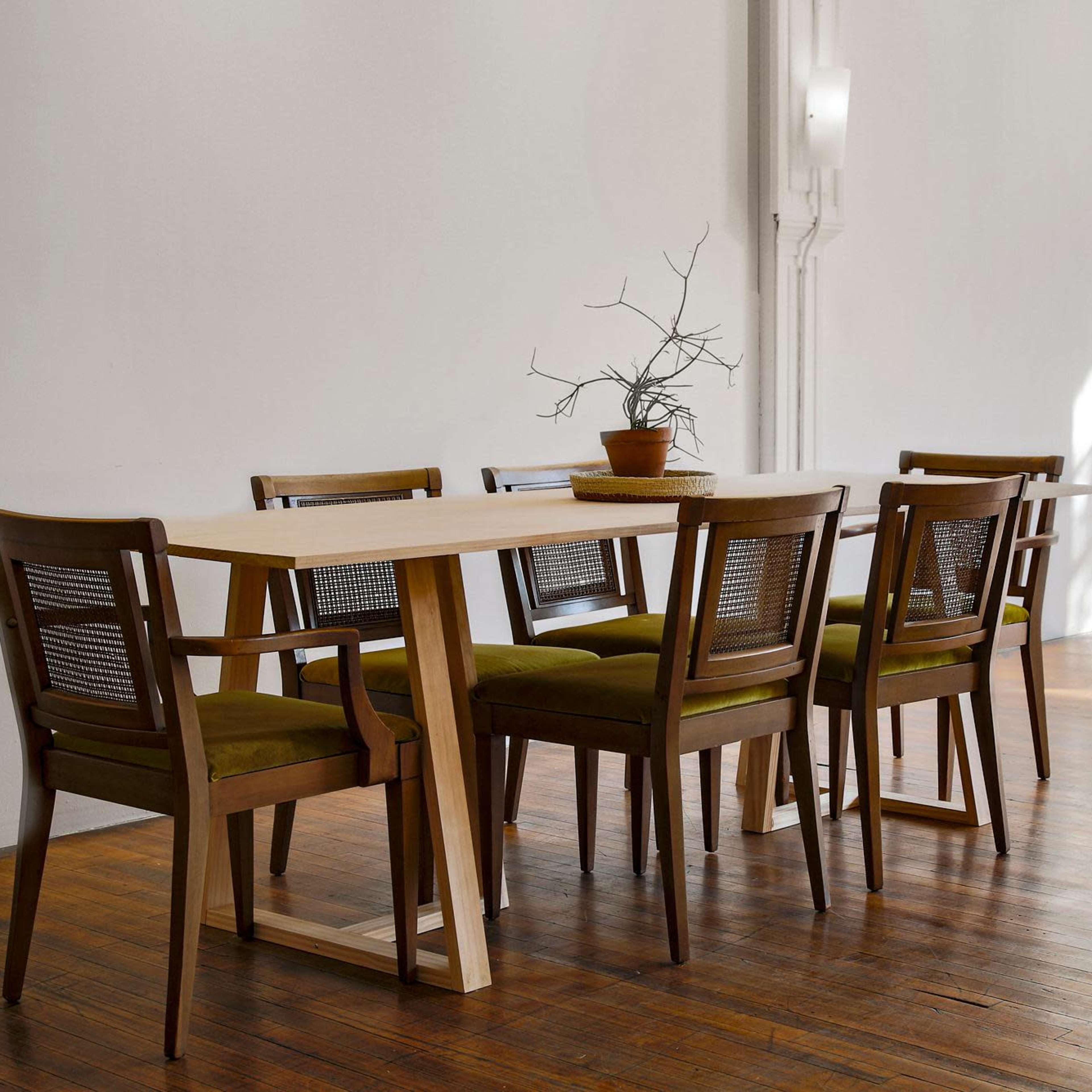 A wooden dining table with six chairs sits in a bright room featuring large windows and a potted plant.