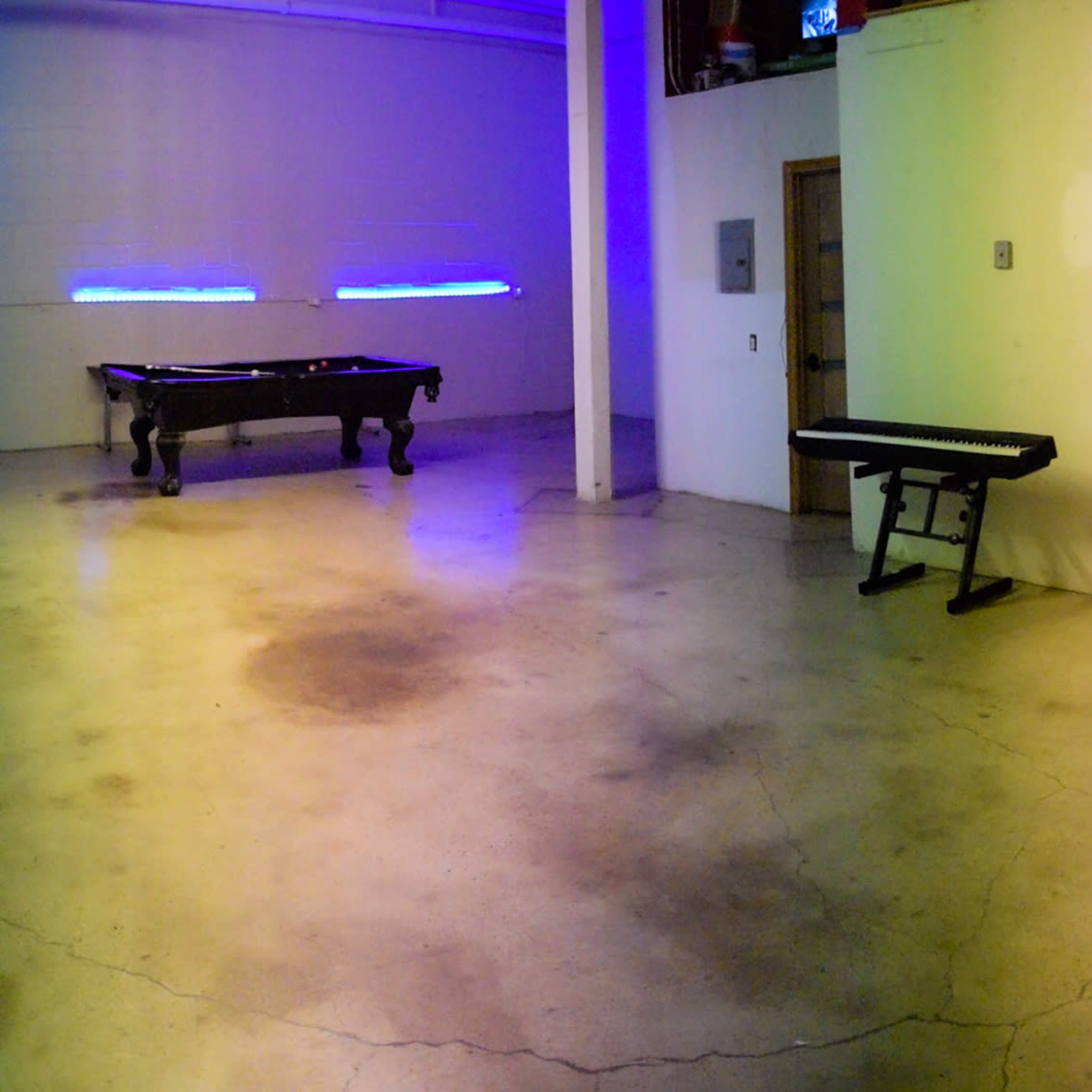 A spacious interior with a pool table, a keyboard, a long table, and a glass-walled room containing musical instruments, all under purple and blue lighting.