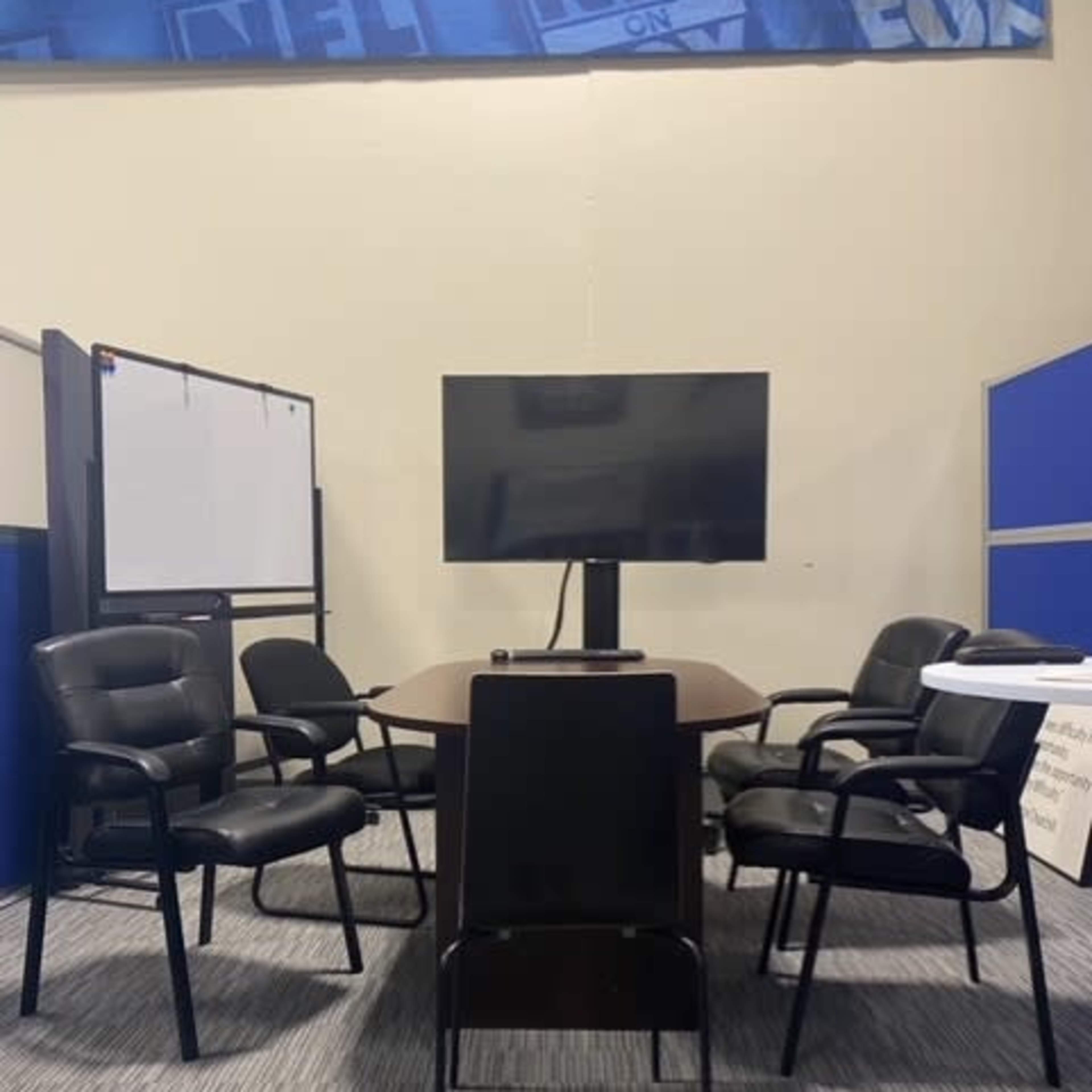 A conference room features a central table surrounded by several black chairs and a mounted television on a stand.
