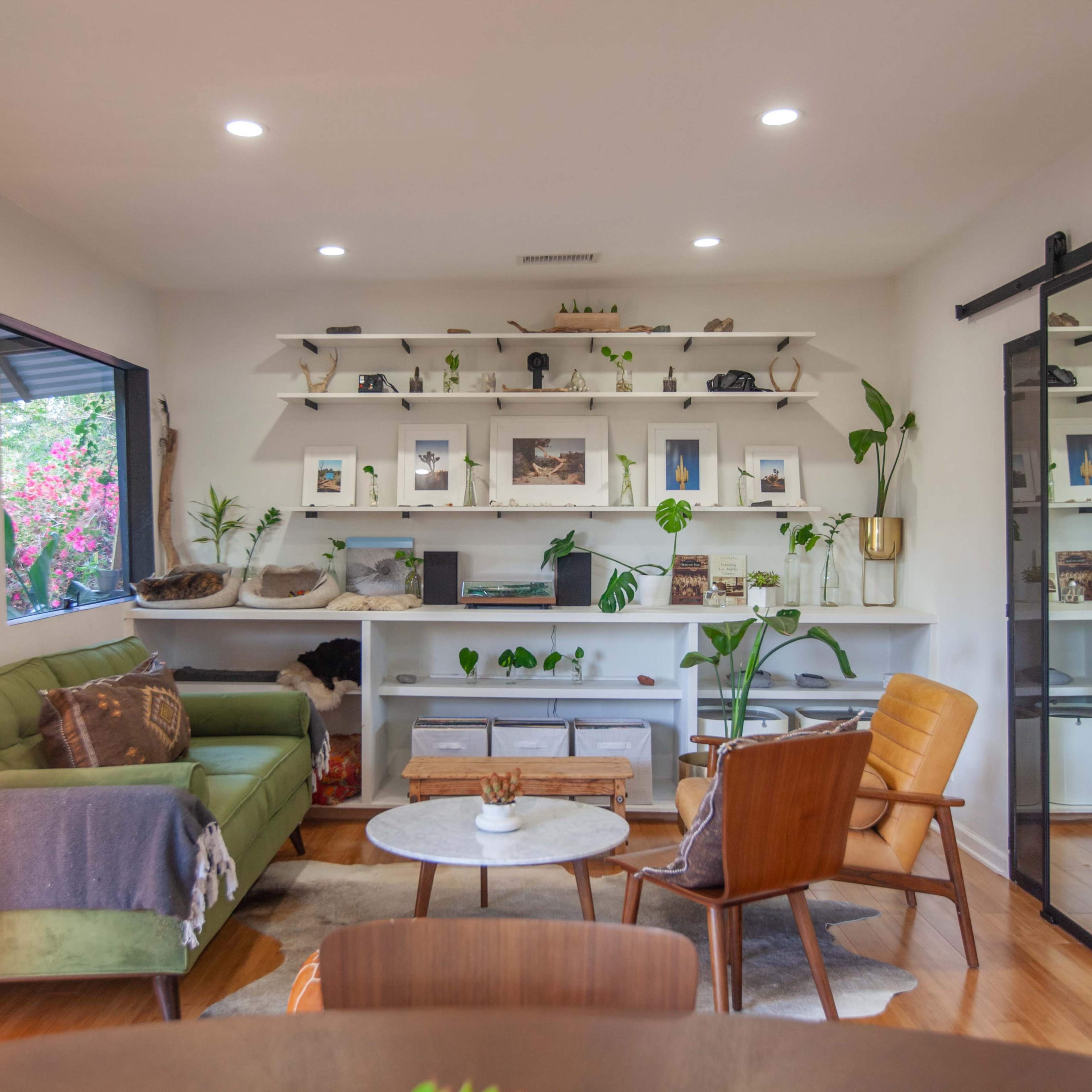 A modern living room features a green sofa, a circular coffee table, and shelves displaying decorative items and plants, with a sliding glass door at one side.
