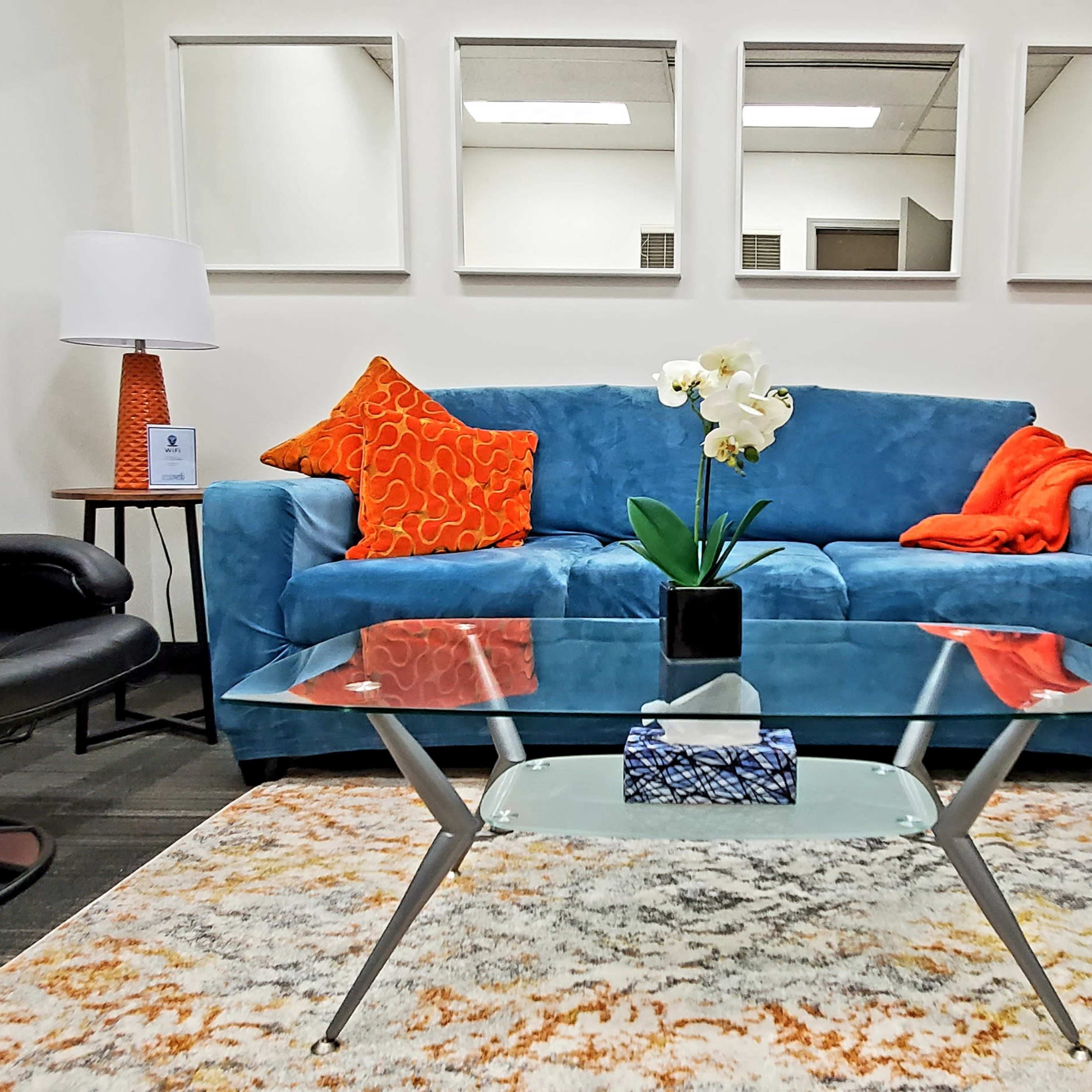 A modern seating area with a blue sofa, two black armchairs, a glass coffee table, and decorative cushions, surrounded by mirrors on the wall.