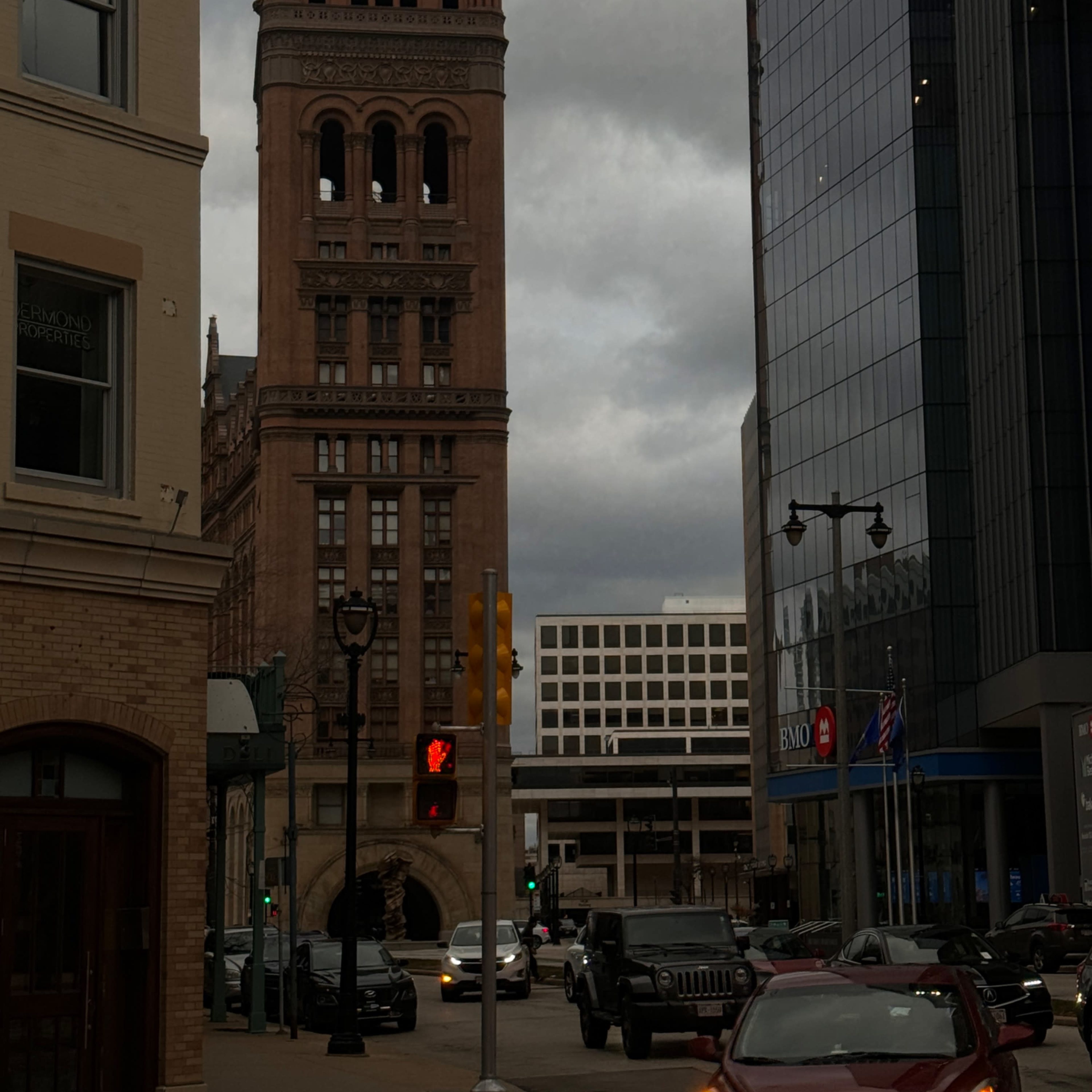 A tall clock tower stands at the end of a city street, framed by modern buildings and overcast skies.