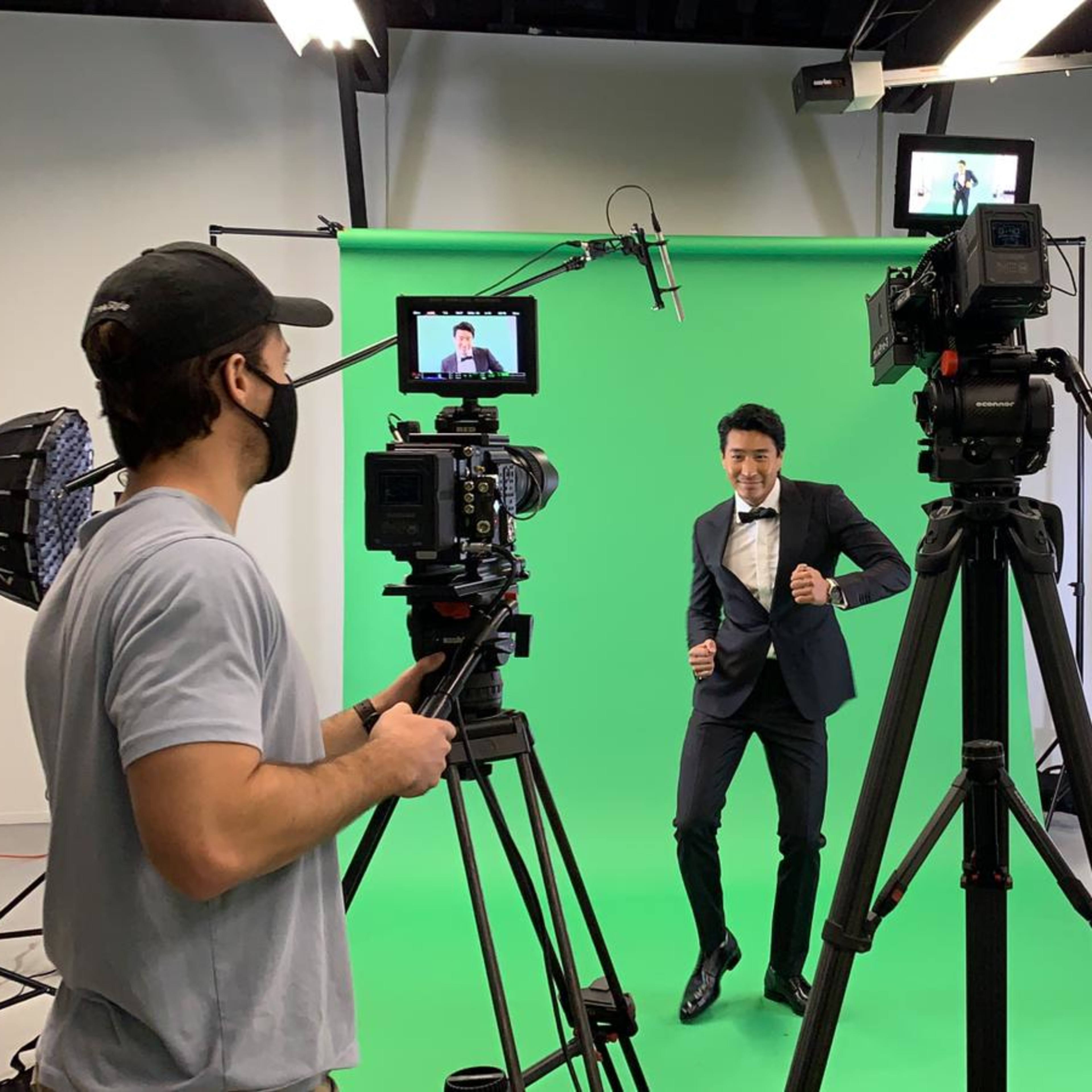 A man in a suit dances in front of a green screen while a camera operator captures the scene with multiple cameras and lighting equipment arranged around them.