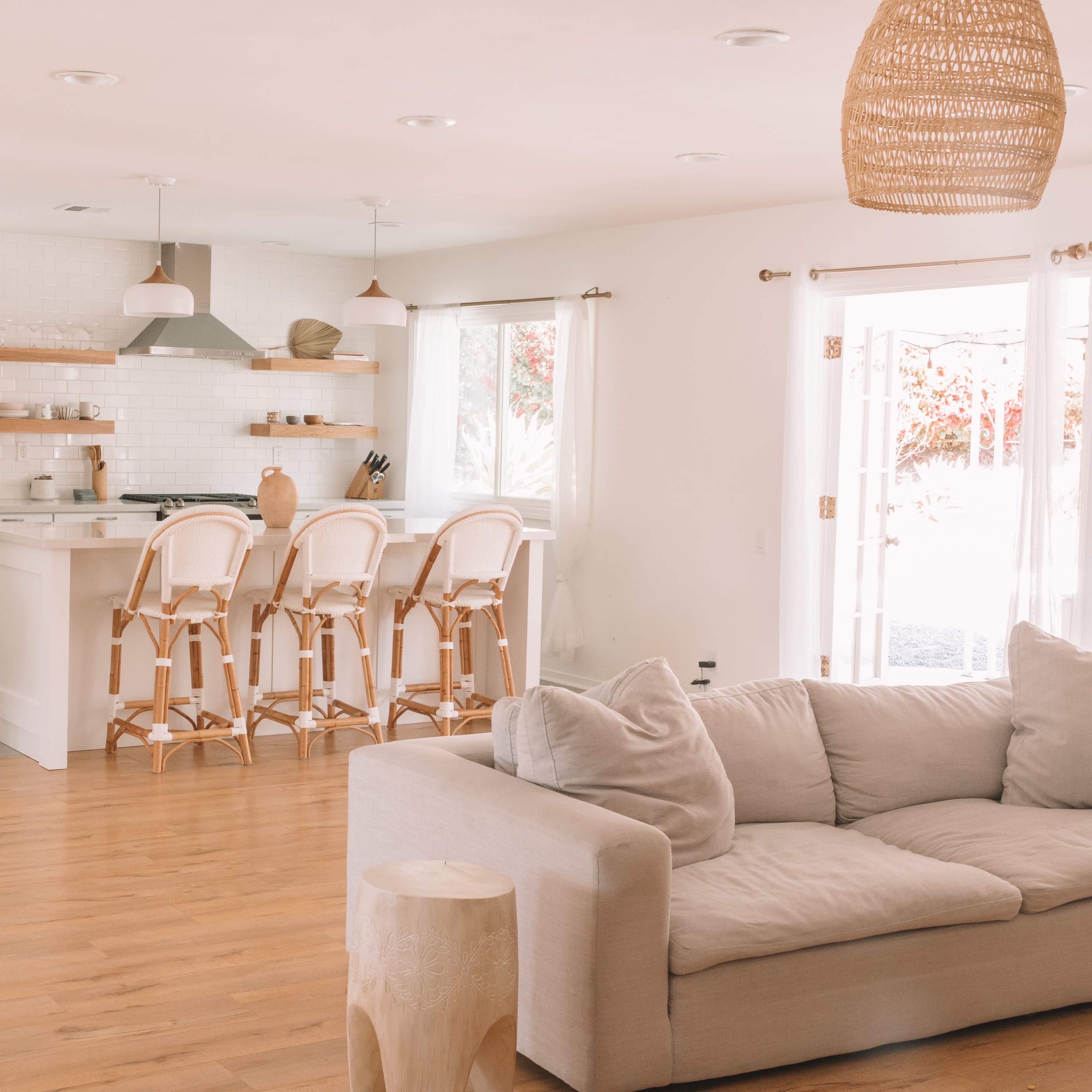 A light-filled open-concept living space with a sofa, a wooden side table, and a kitchen area featuring bar stools and shelves.