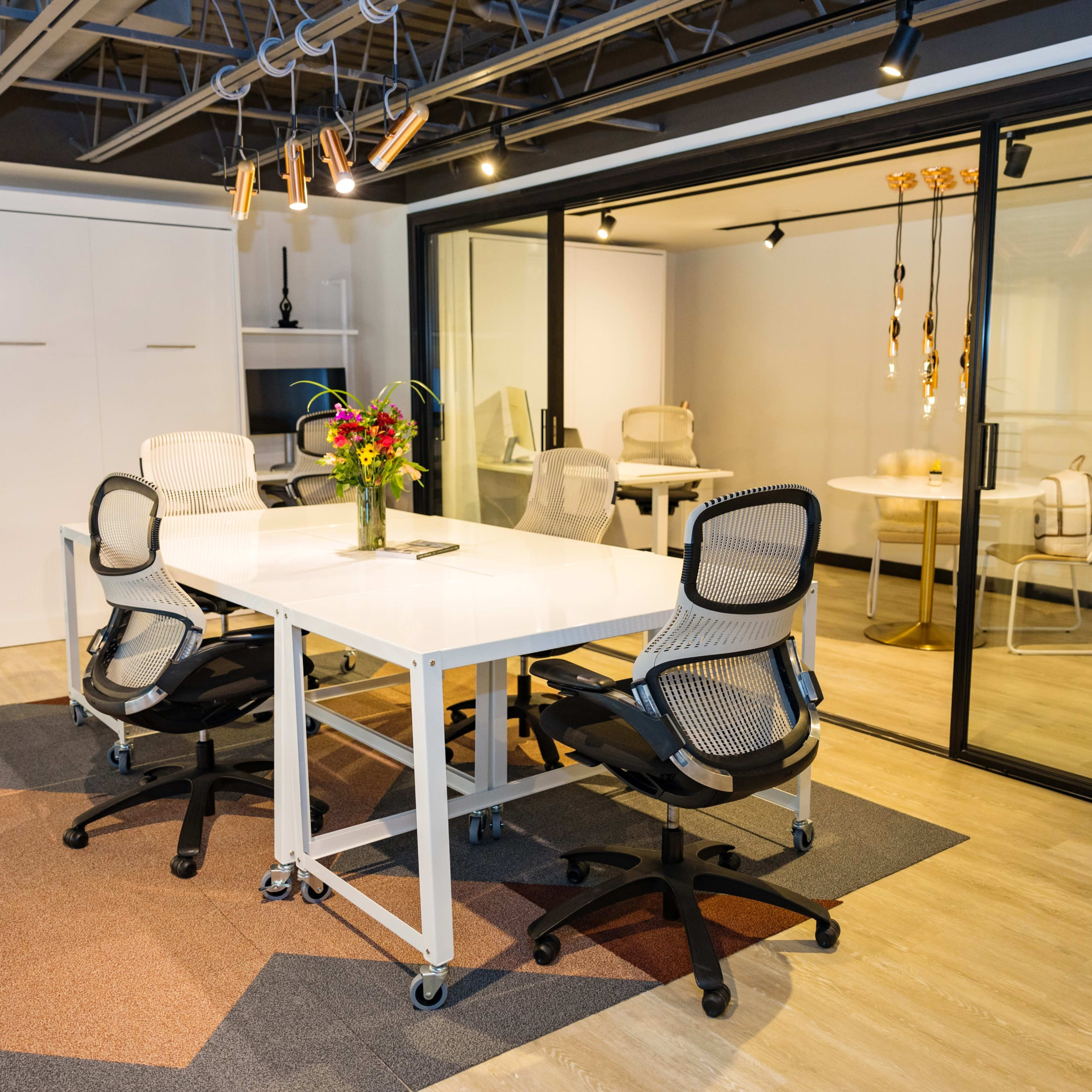 The scene shows a modern office space with a central white table surrounded by rolling office chairs, and a glass-walled conference room in the background.