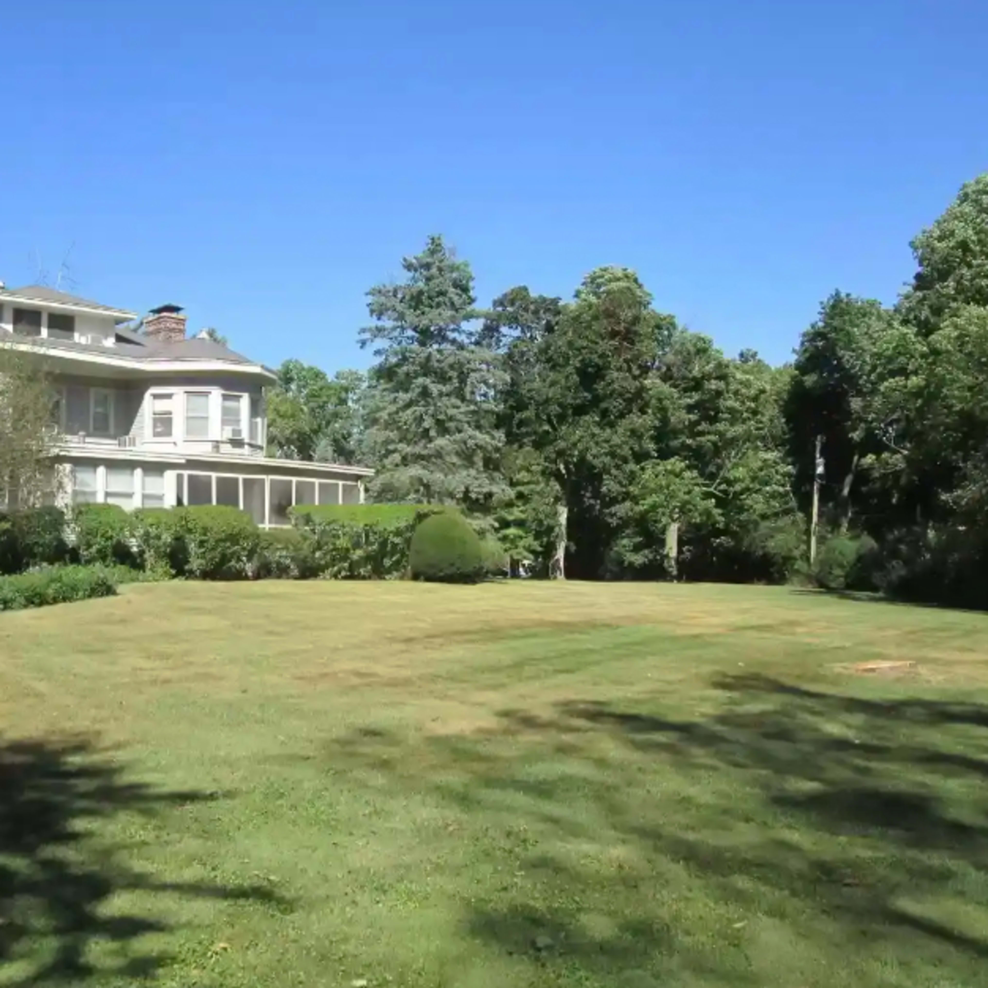 A large house with multiple stories is set beside a grassy lawn surrounded by trees under a clear blue sky.