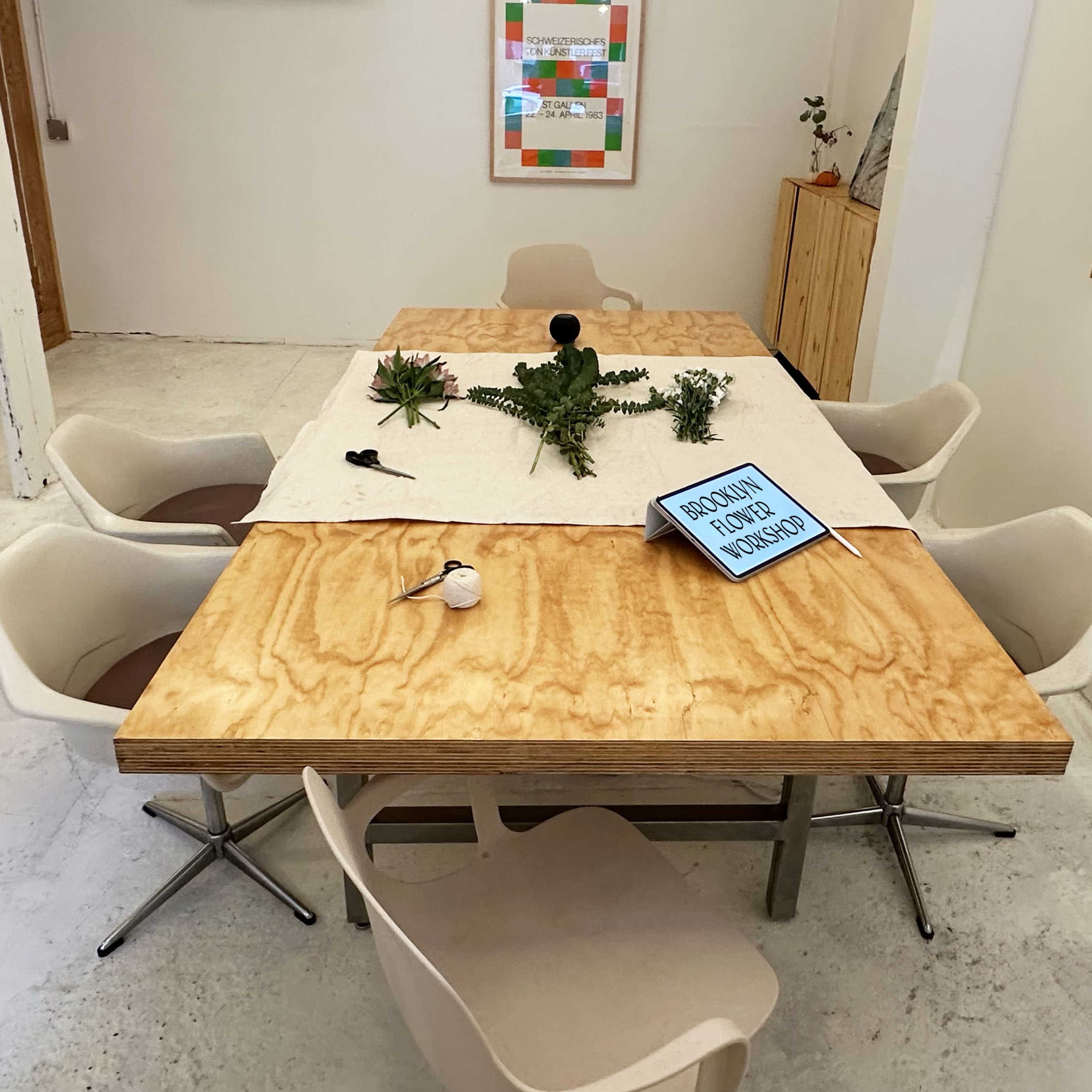 A wooden table with a beige cloth on top is surrounded by six chairs, with various plants and a sign displayed on the table in a well-lit room.