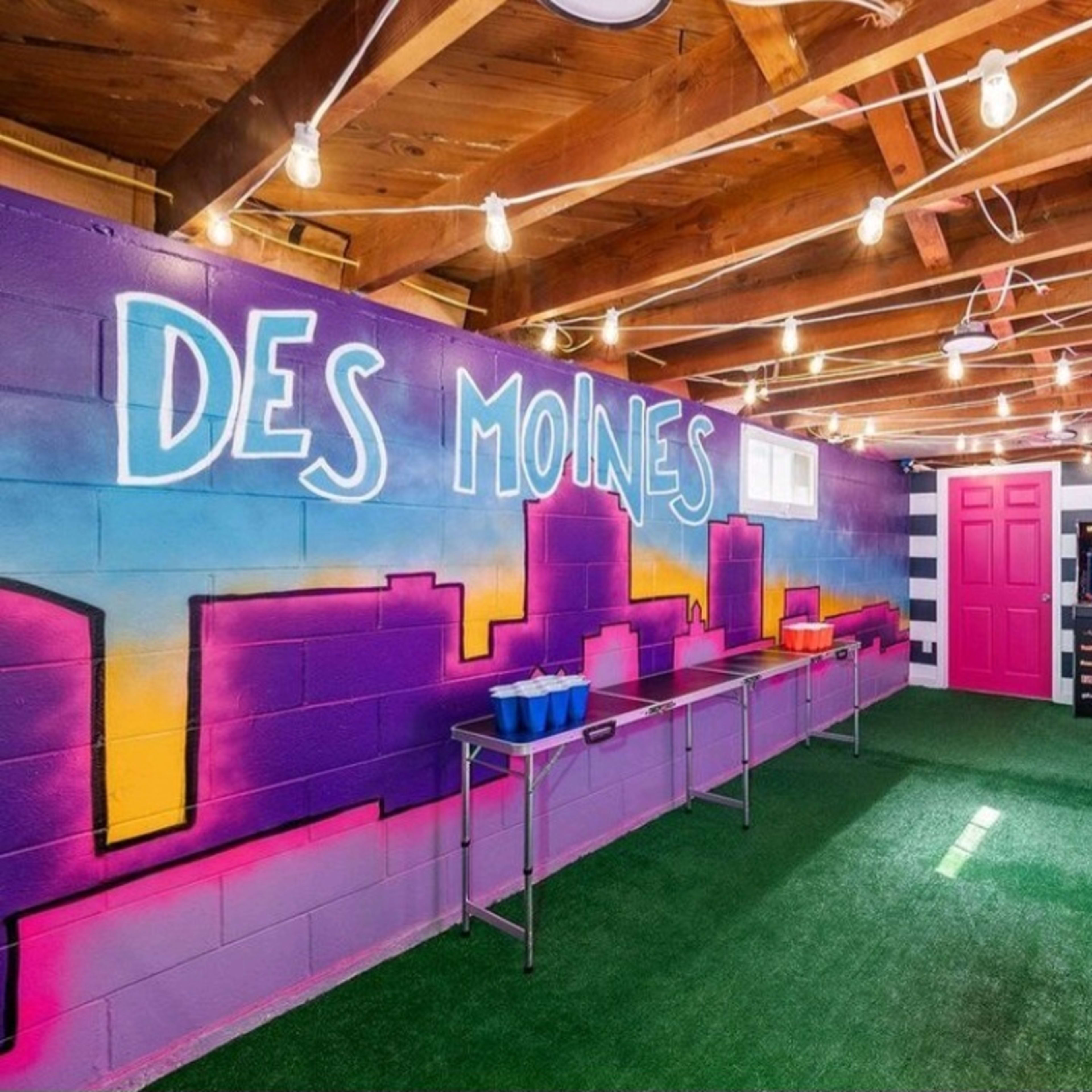 A brightly colored indoor space with purple and pink walls, artificial grass flooring, and string lights, featuring tables and a vending machine.