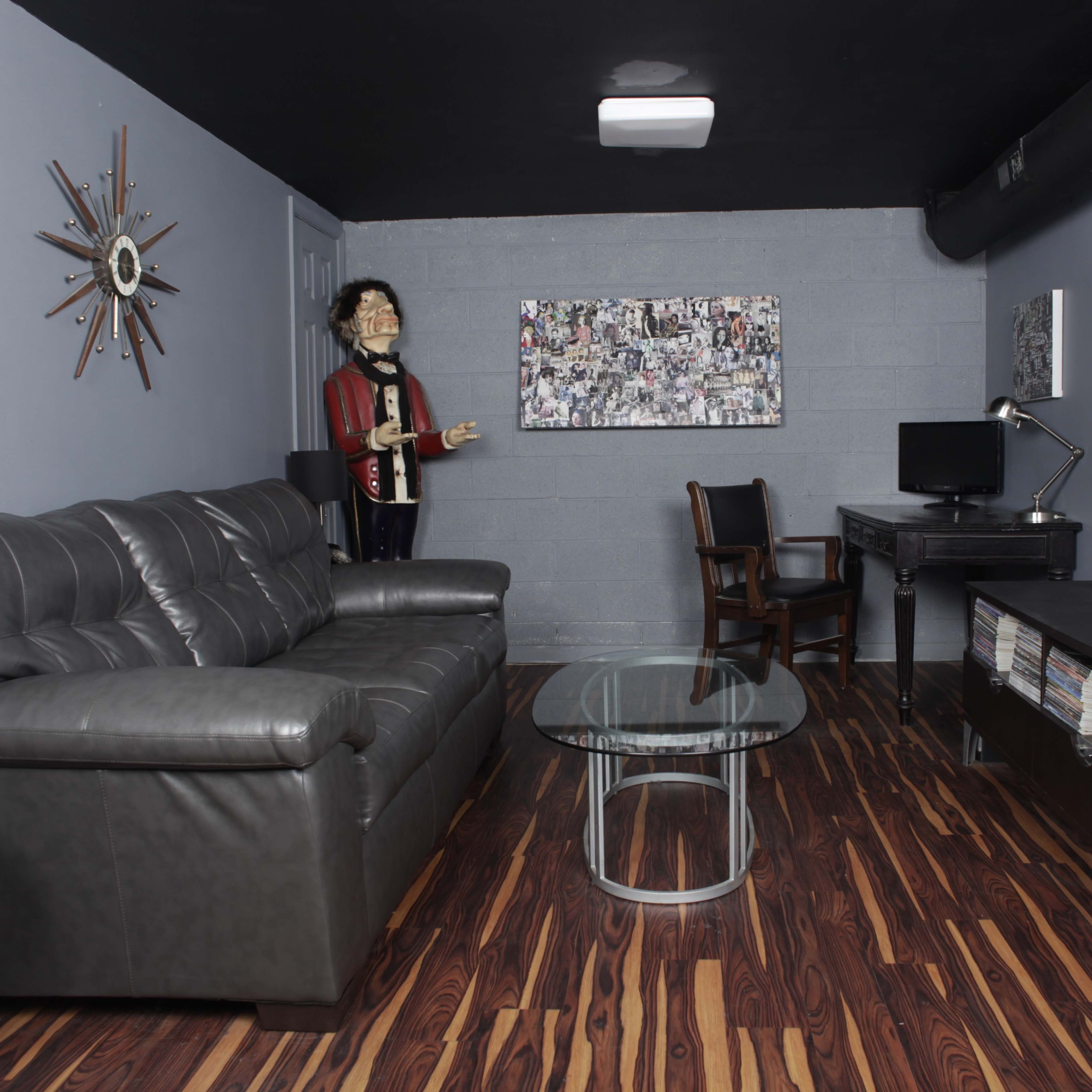 The room features a gray wall, a black leather couch, a glass coffee table, a wooden chair, a desk with a computer, and a life-sized mannequin dressed in a suit.