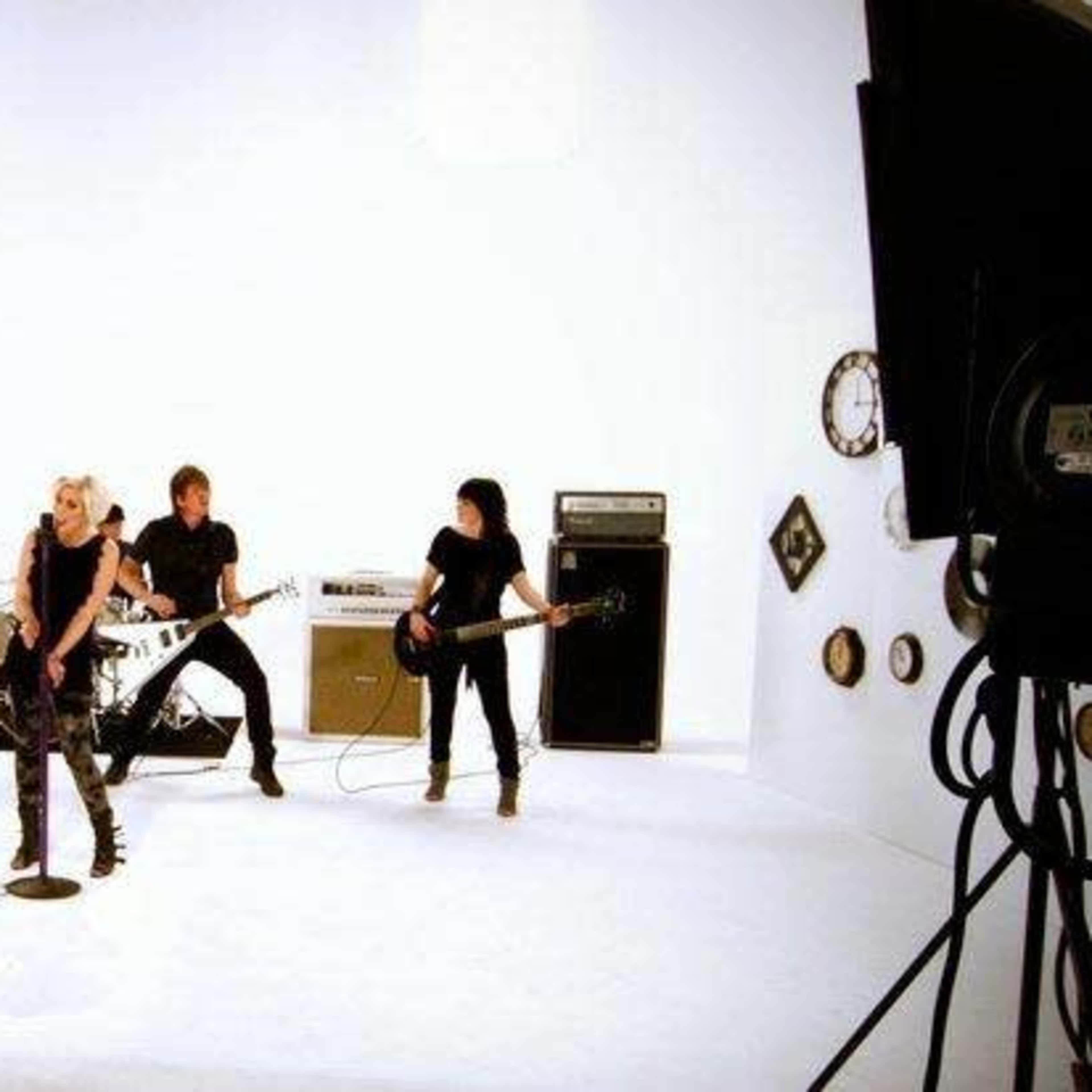 A band performs on a white set while a camera crew films the scene.