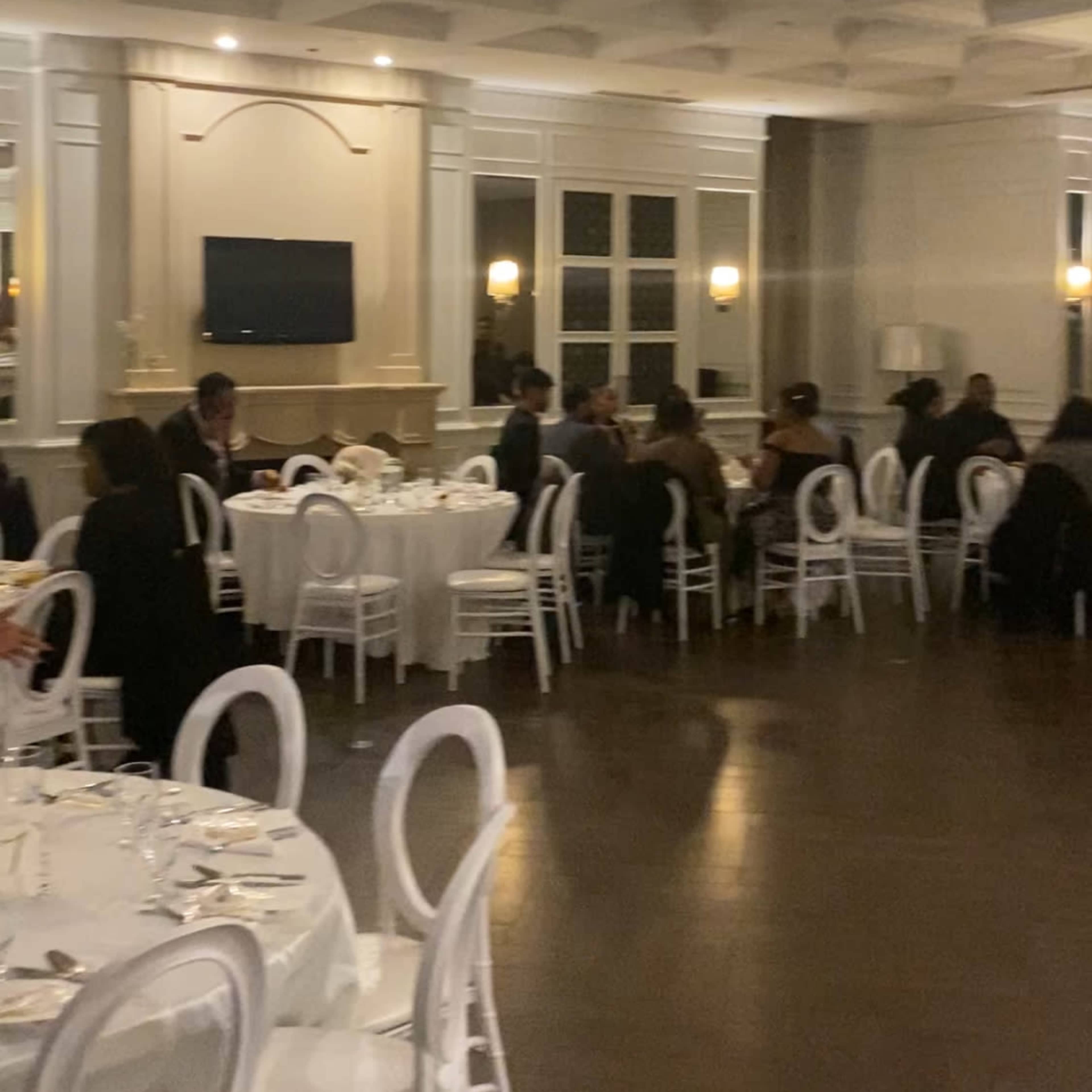 A spacious dining room features round tables set for a formal event, with guests seated and engaged in conversation.