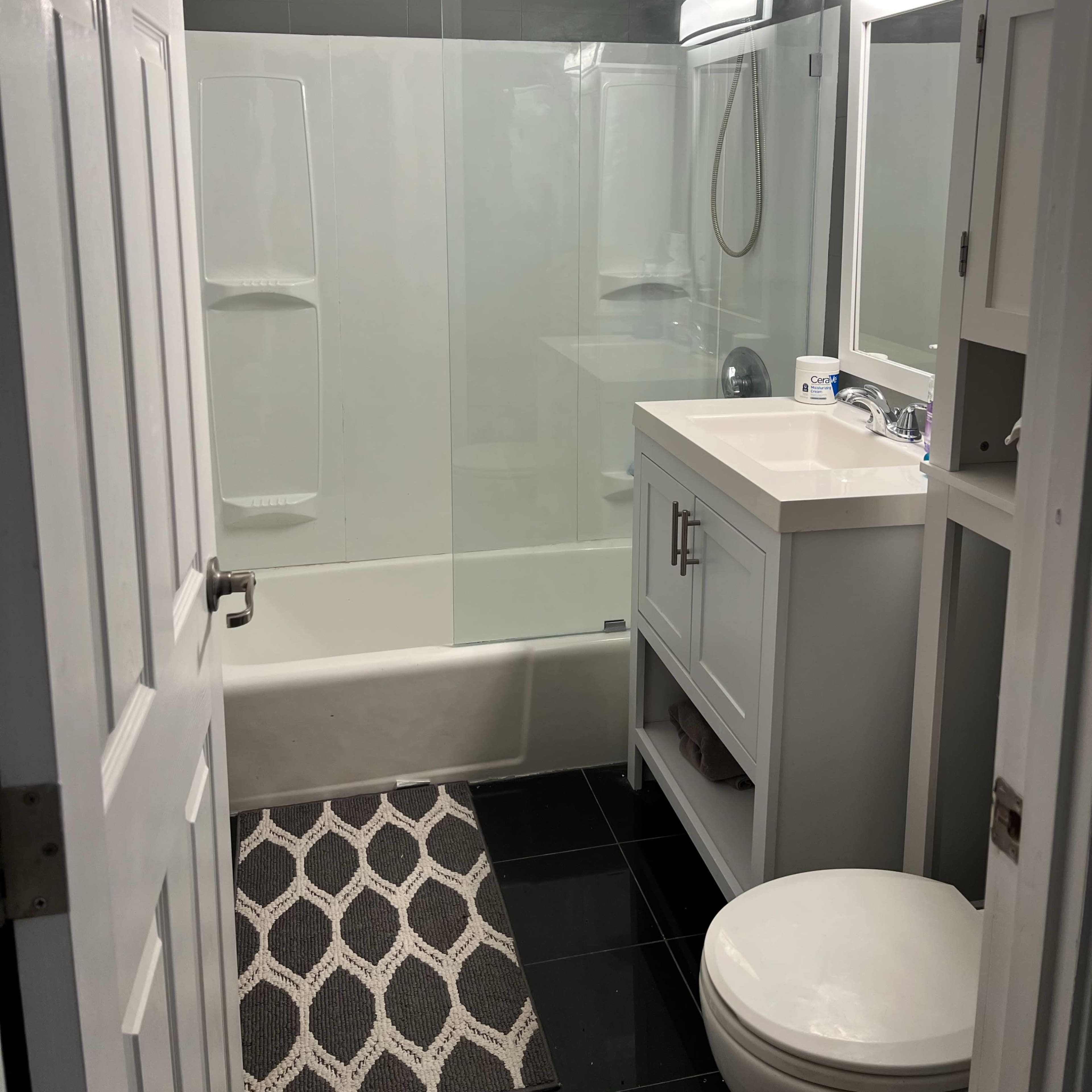 A modern bathroom featuring a glass shower enclosure, a white vanity, a toilet, and a geometric patterned rug on the black tile floor.