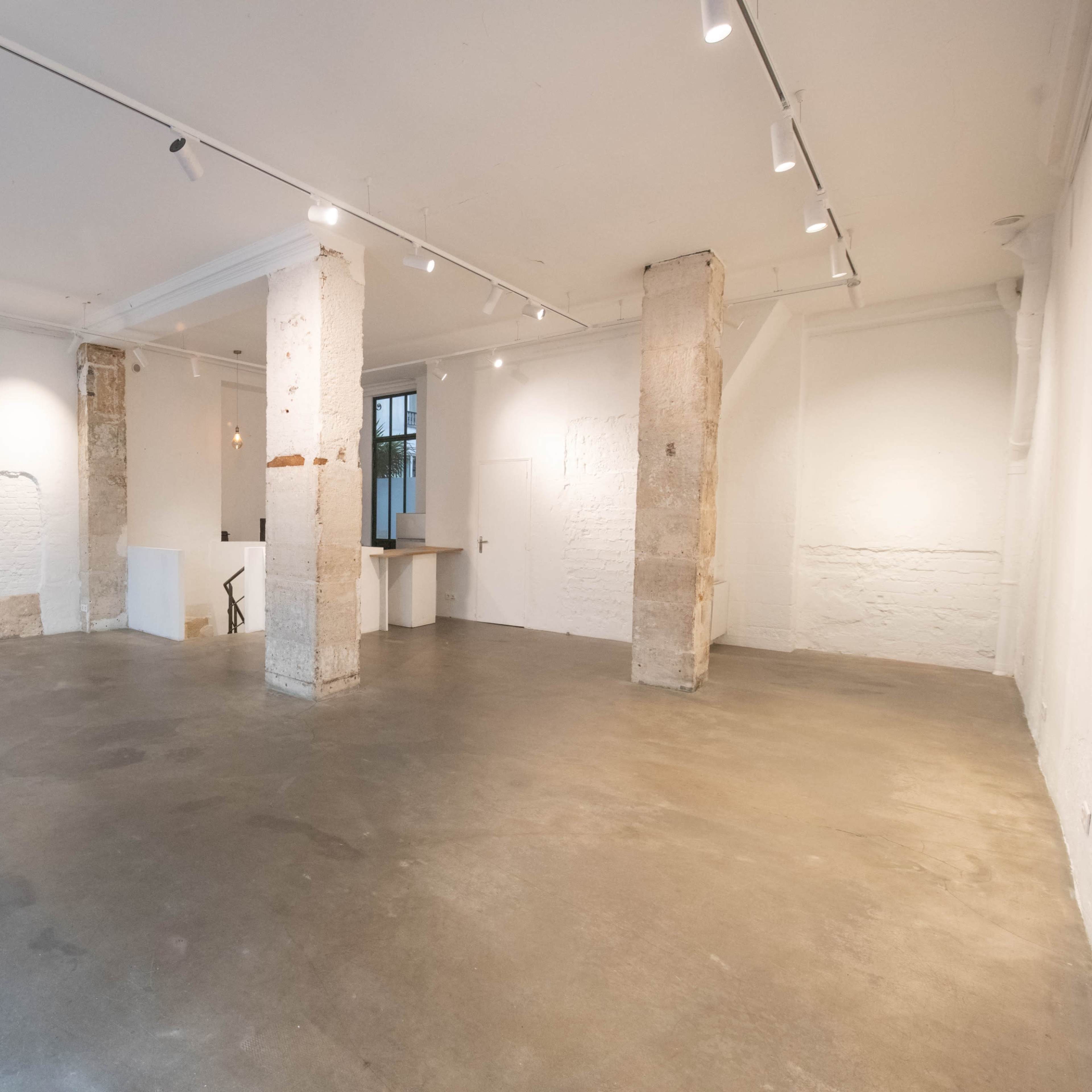 A spacious, minimalist gallery interior with concrete floors and exposed pillars, featuring white walls and soft overhead lighting.