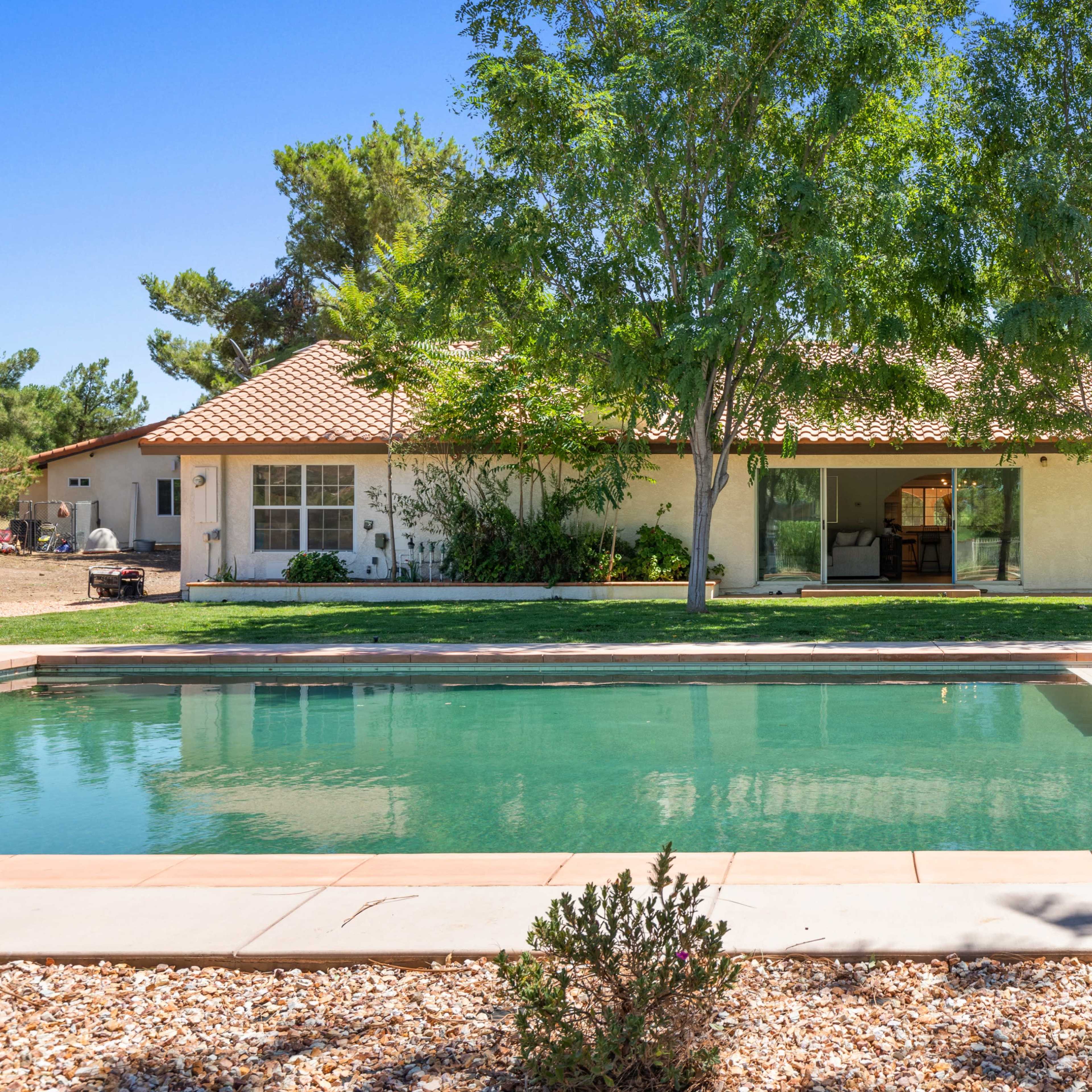 A house with a tile roof adjacent to a rectangular swimming pool surrounded by trees and landscaping.