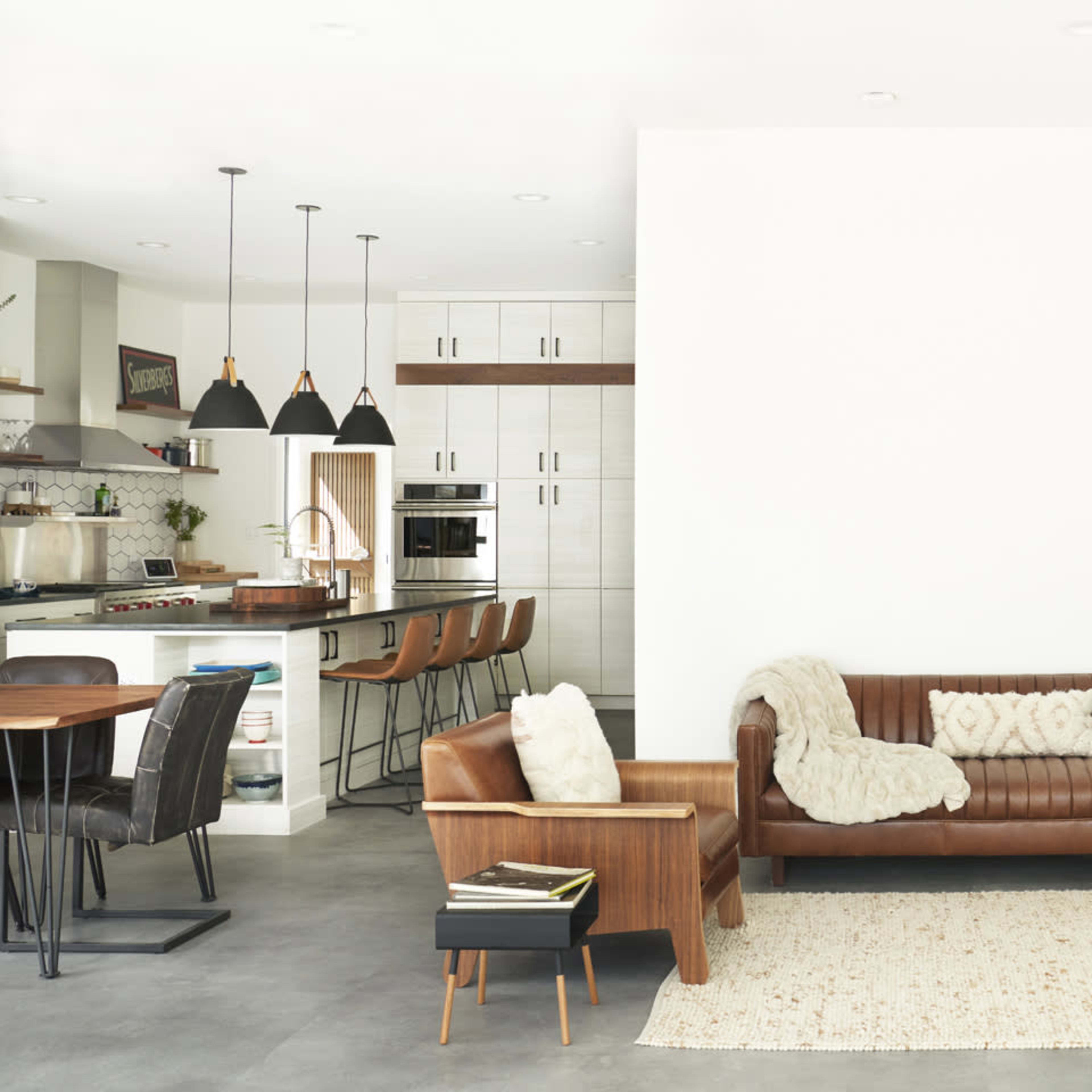 A modern living room features a brown leather sofa, a wooden armchair, a white wall, and a flat-screen TV, with an open kitchen and dining area in the background.