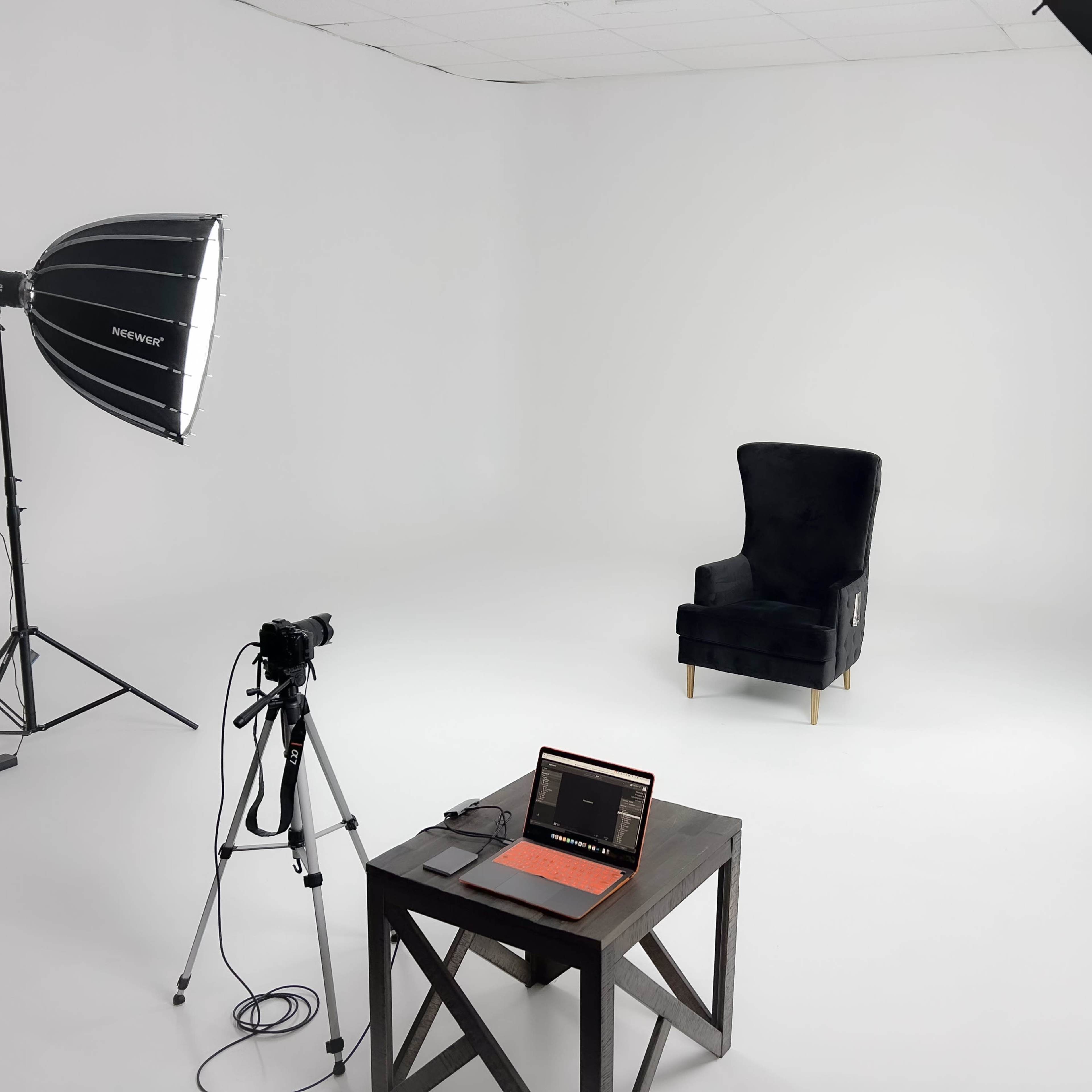 A minimalist studio setup with a black velvet chair, a camera on a tripod, and a laptop on a wooden table, illuminated by two softbox lights.