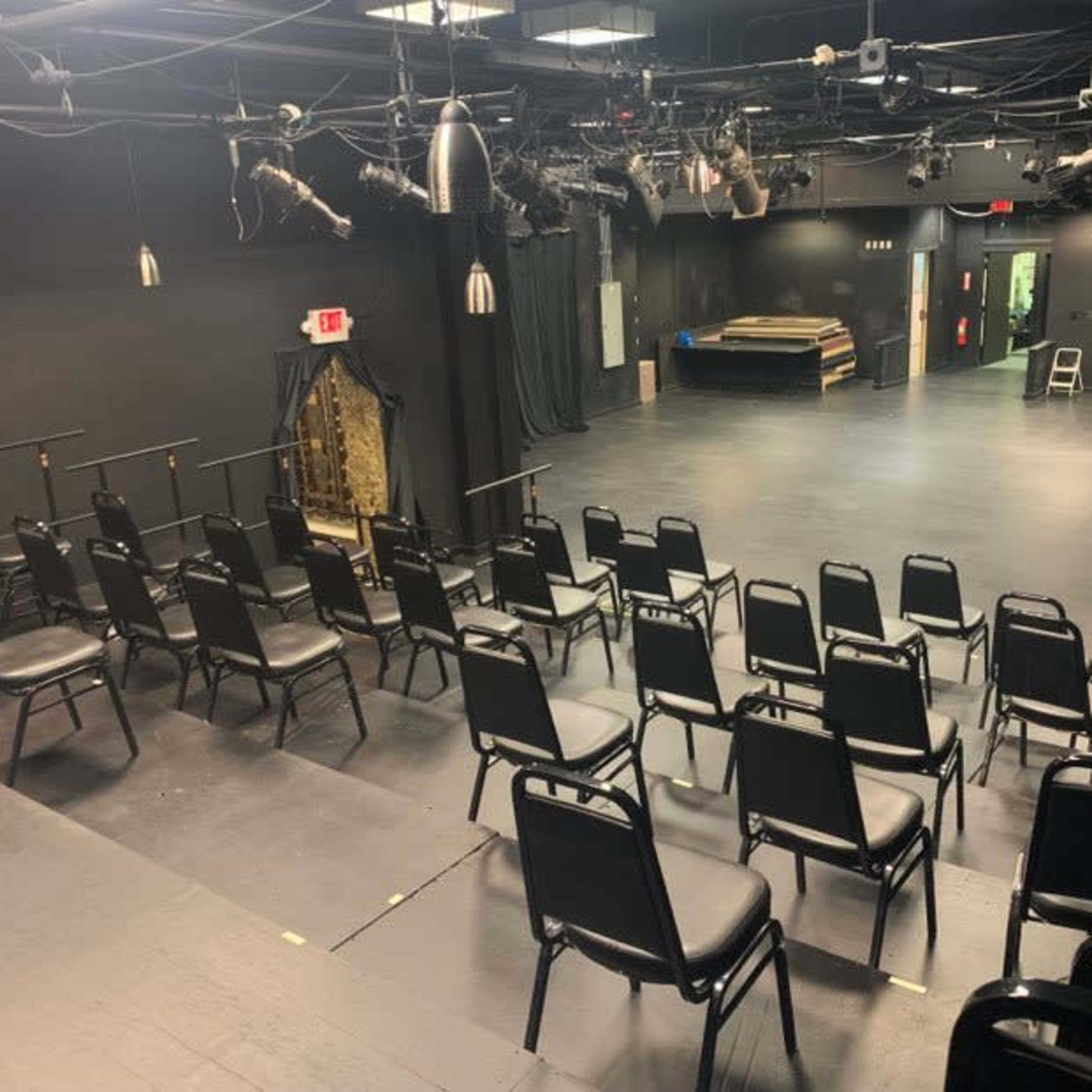 A small black box theater with rows of black chairs facing a performance space illuminated by overhead stage lights.