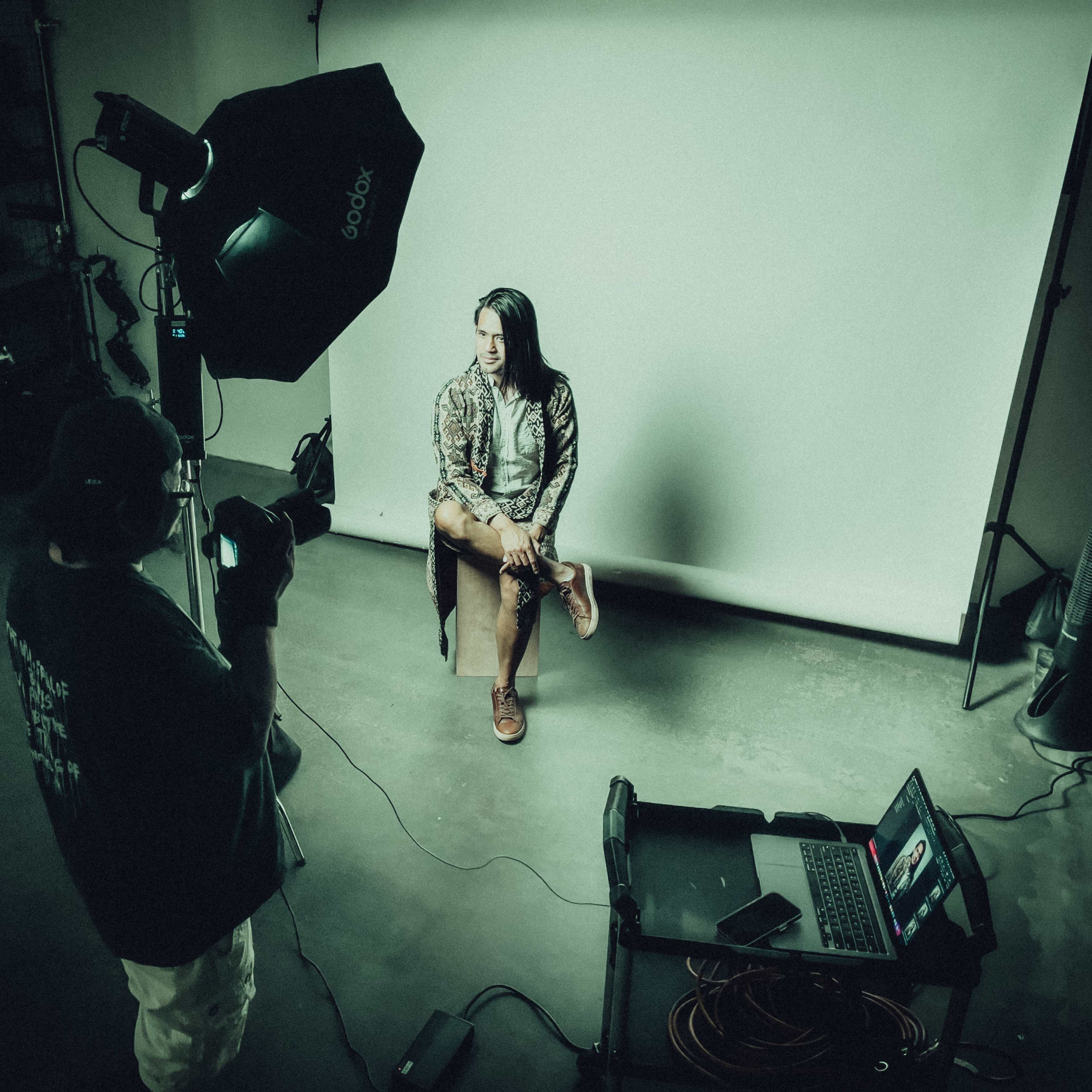 A photographer is capturing a portrait of a person sitting on a stool in a studio with a backdrop and lighting setup.
