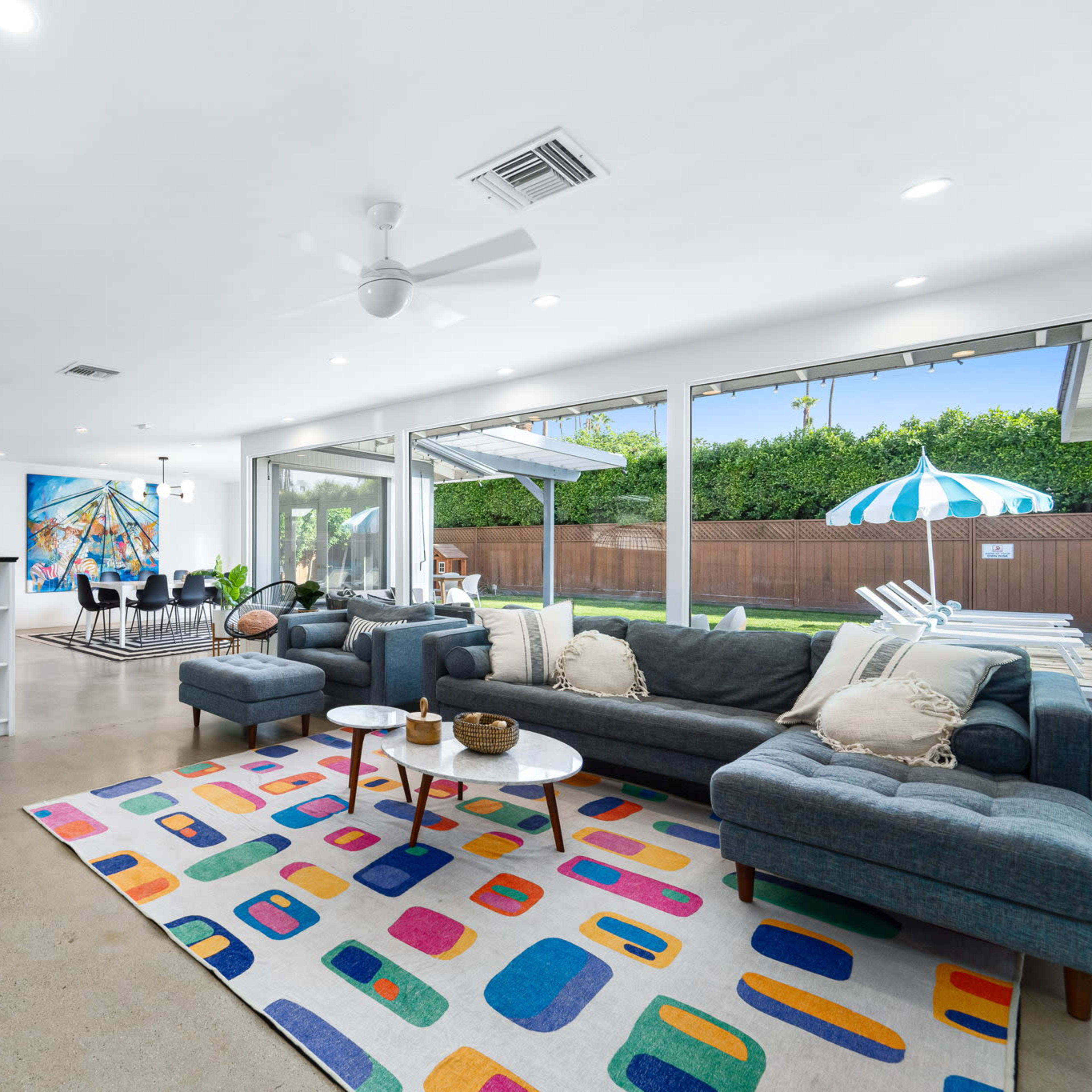 A spacious living area features a gray sectional sofa, vibrant area rug, and large windows overlooking a patio with a swimming pool.