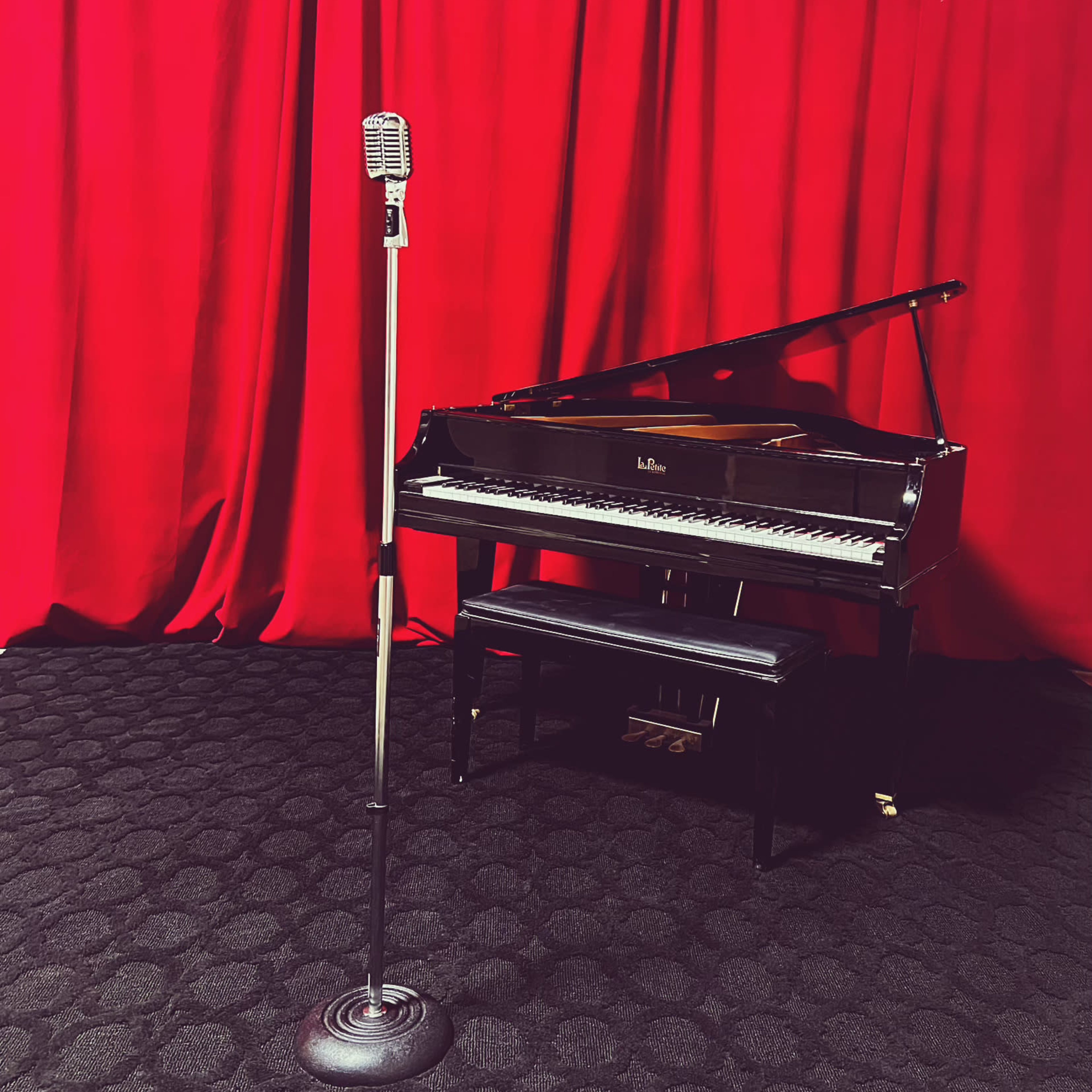 A vintage microphone stands next to a grand piano set against a red curtain backdrop.
