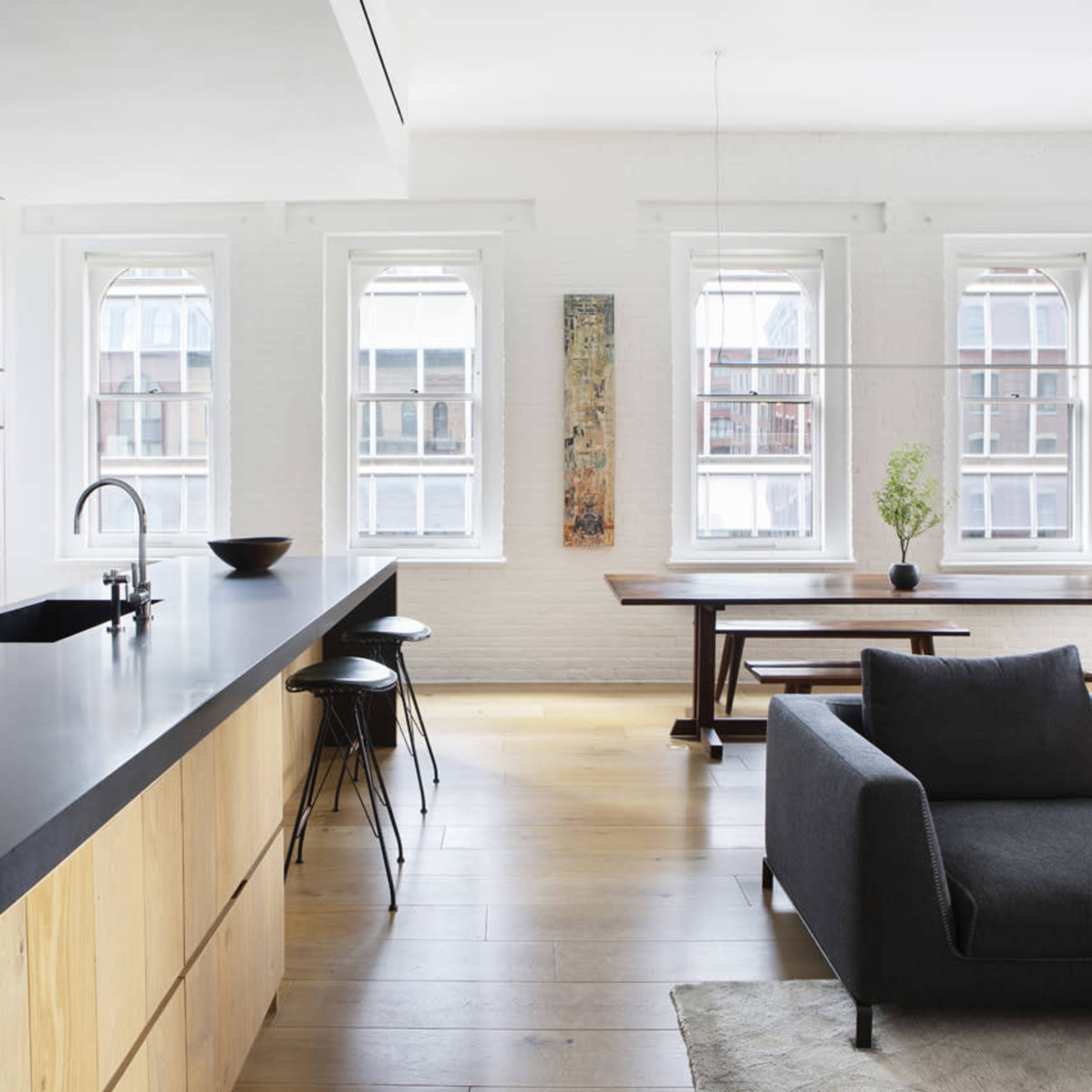 A modern kitchen and living area featuring a long black countertop, wooden cabinets, and large windows that illuminate the space.