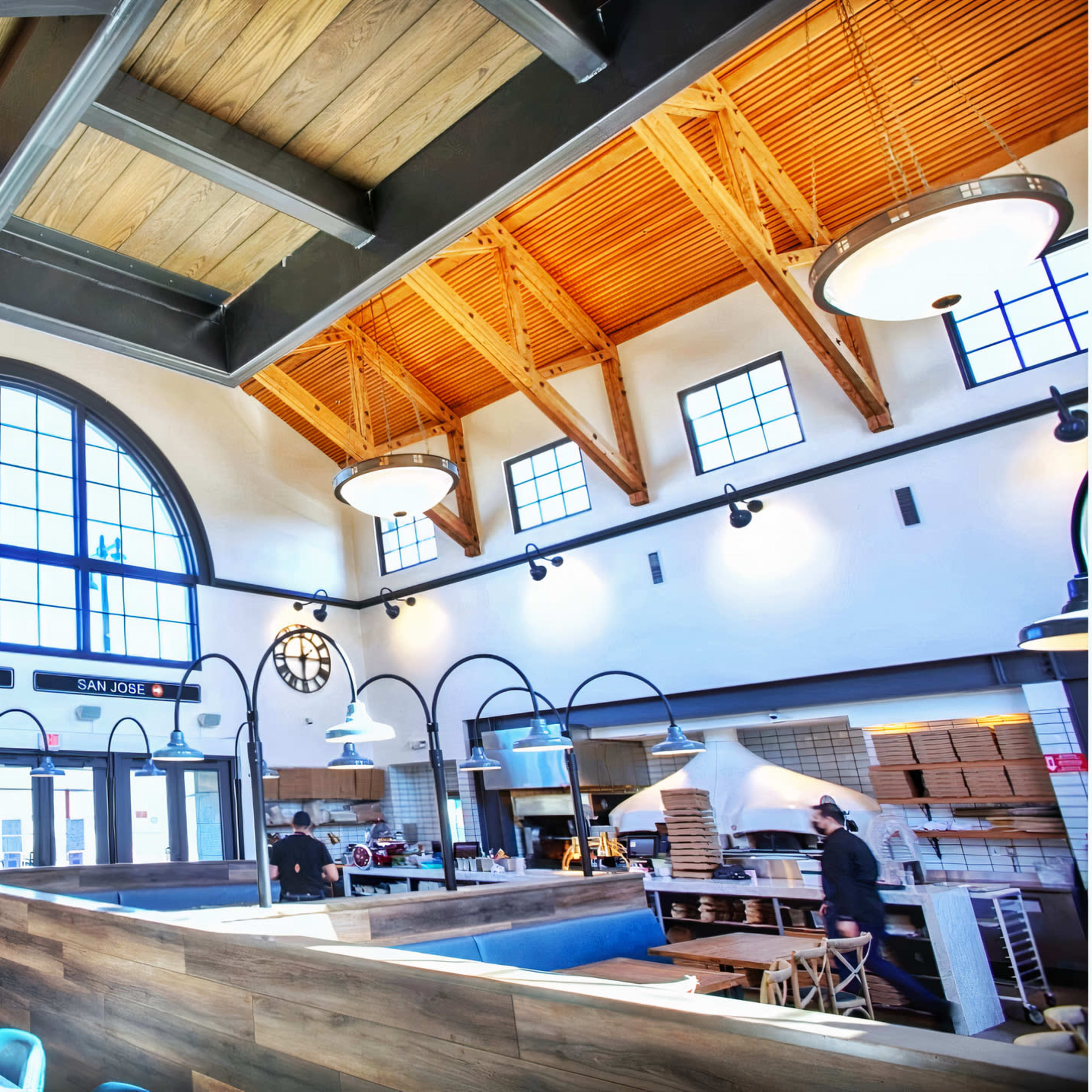 A spacious restaurant interior with a high wooden ceiling, large arched windows, and a long bar area with stools.