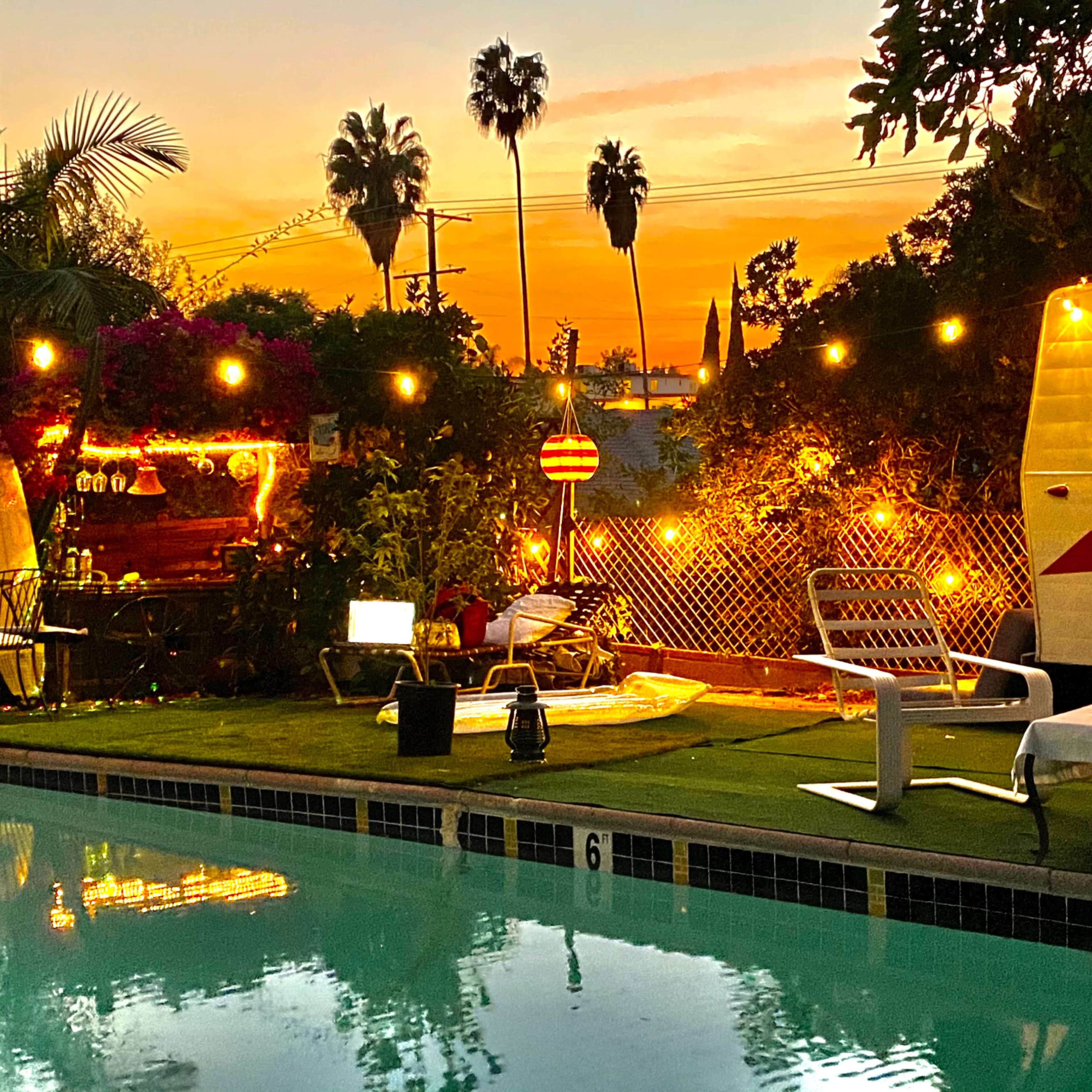 A swimming pool is illuminated by string lights, with a vintage camper, lounge chairs, and tropical plants in the background during sunset.