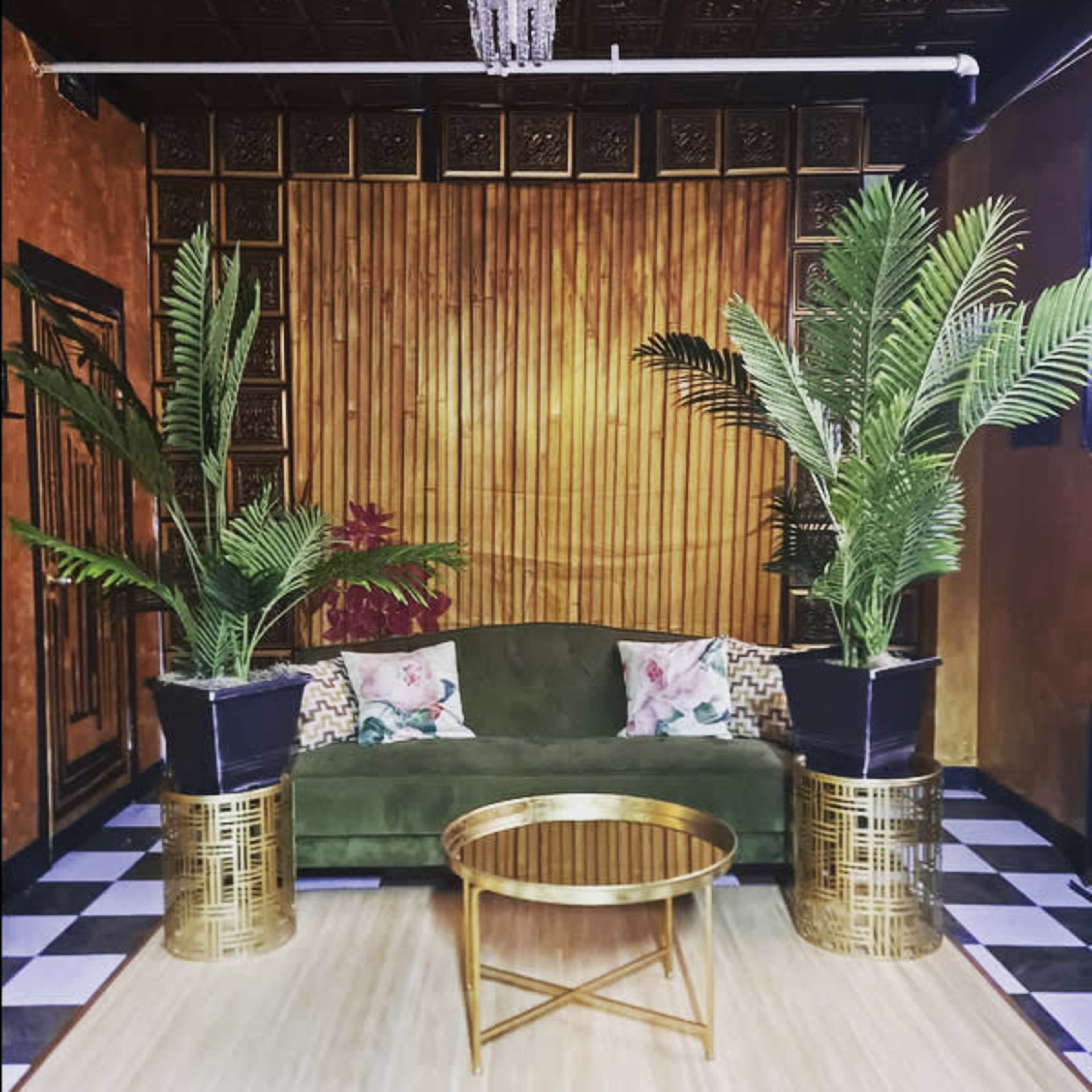 A living room with a green sofa, gold accent tables, and potted palm plants against a backdrop of wooden slats and a patterned ceiling.