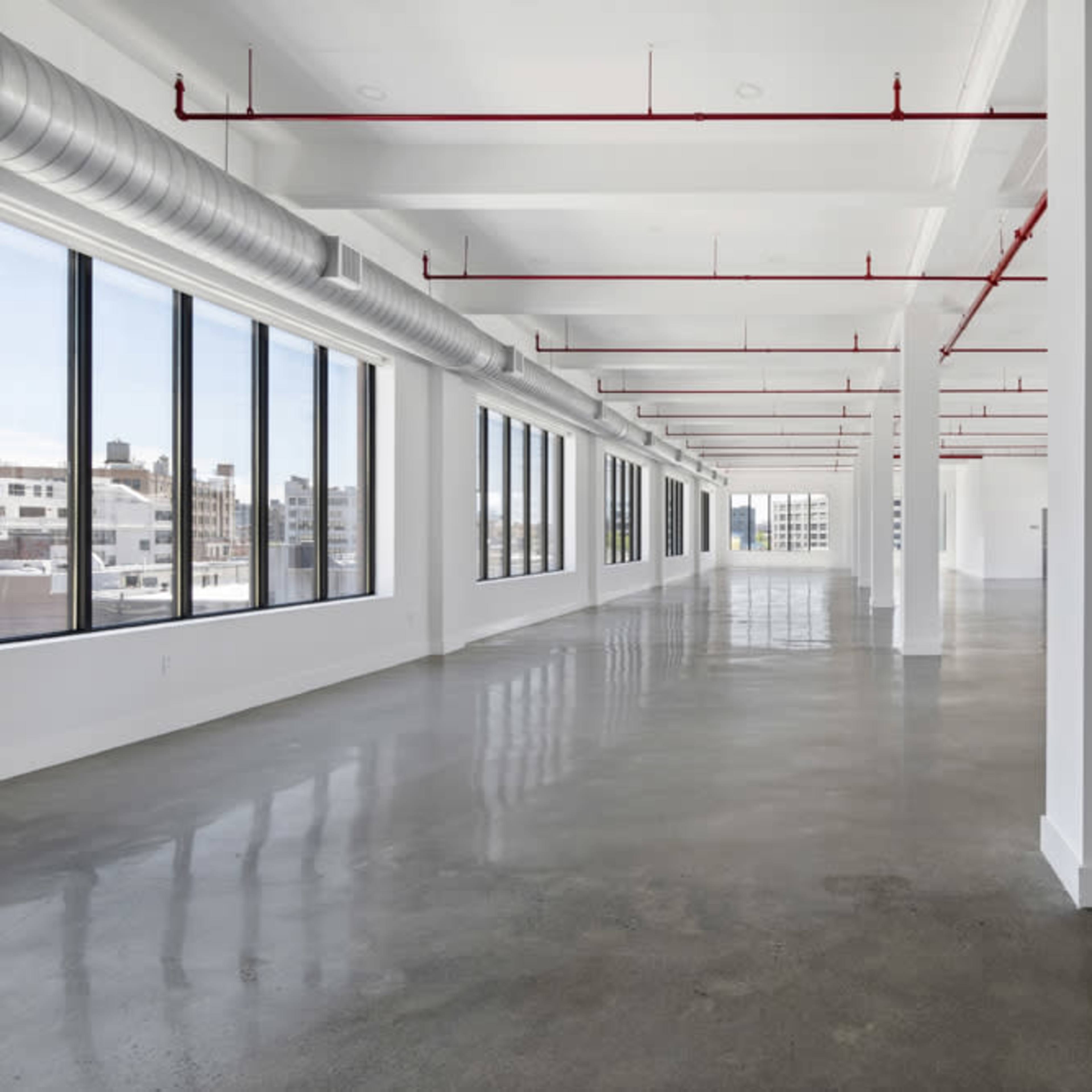 A spacious, unfinished commercial interior with concrete flooring and large windows, featuring exposed red piping along the ceiling.