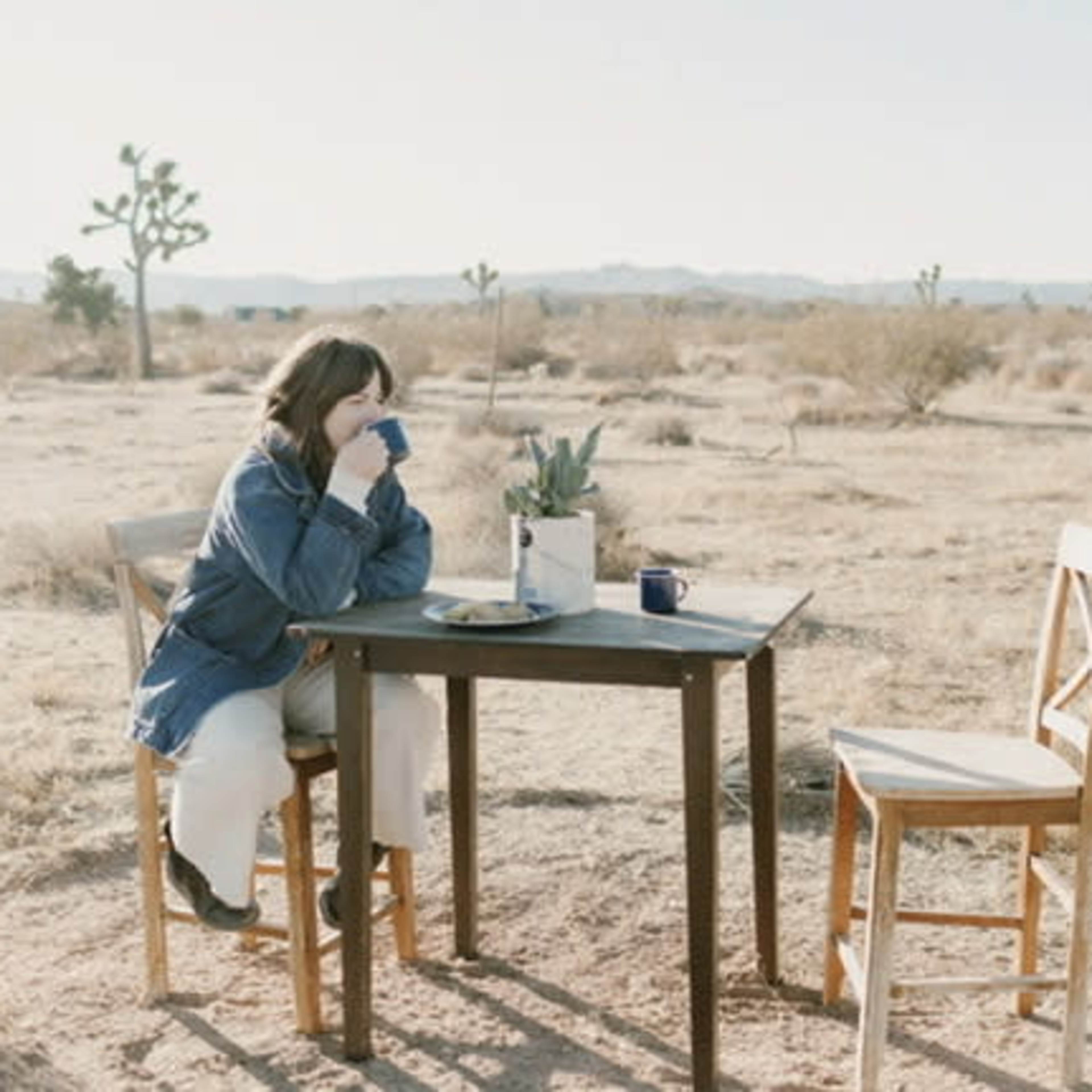A person sits at a table in a desert landscape, holding a blue mug with a few chairs nearby.