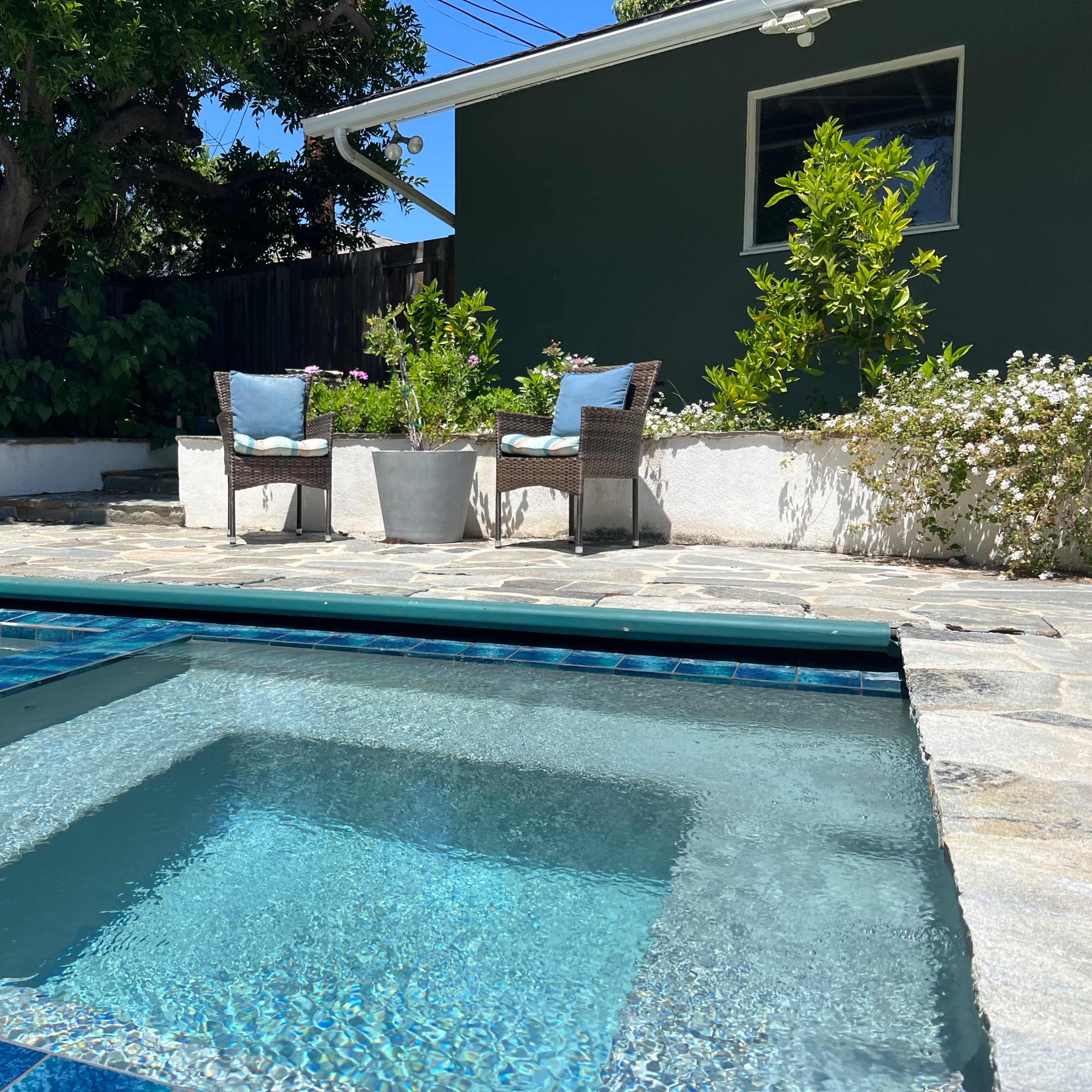 A rectangular pool with a spa section sits beside a landscaped area featuring two wicker chairs and plants.