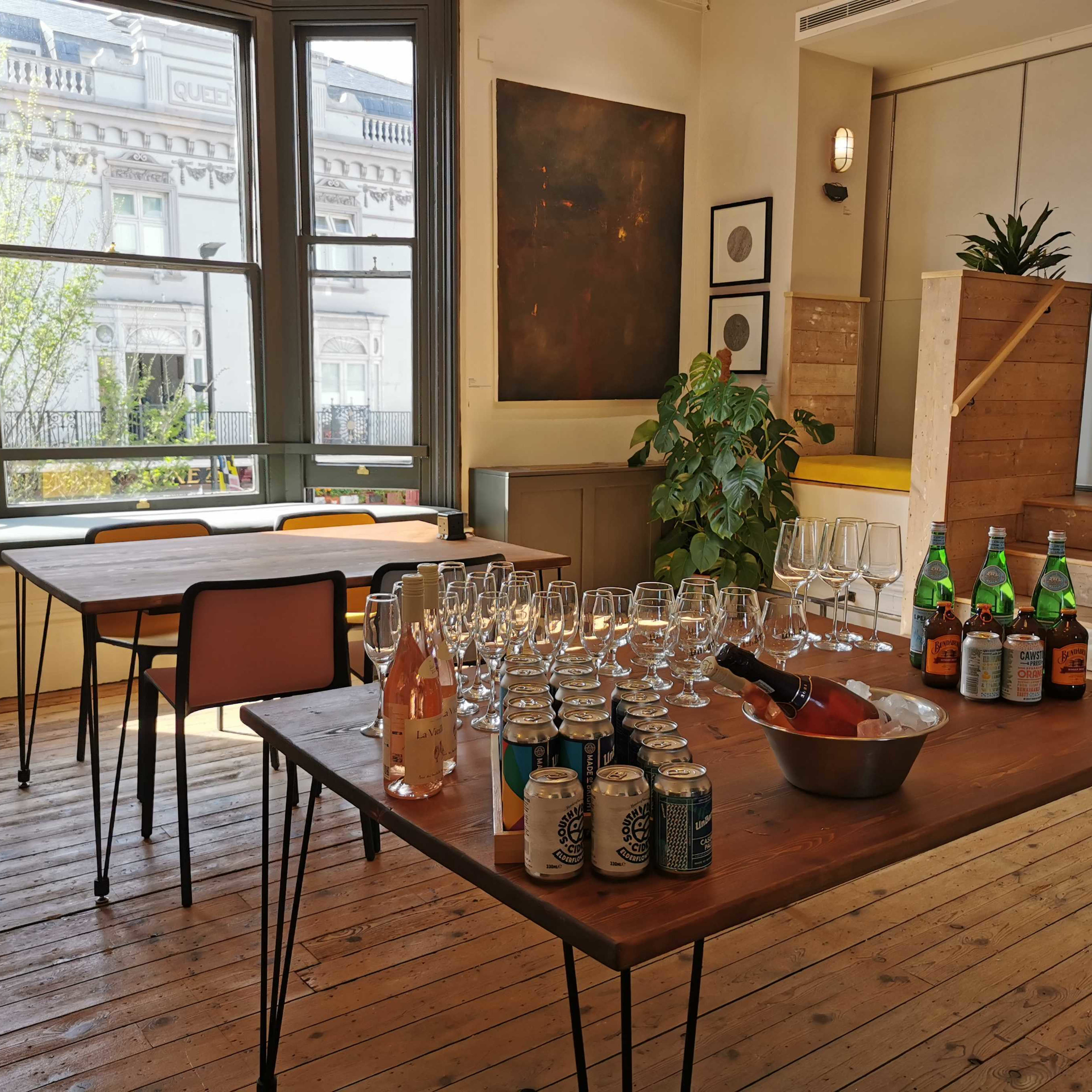 A room features multiple tables with a display of glasses, a bowl of ice with bottles, and several cans arranged on a wooden surface.