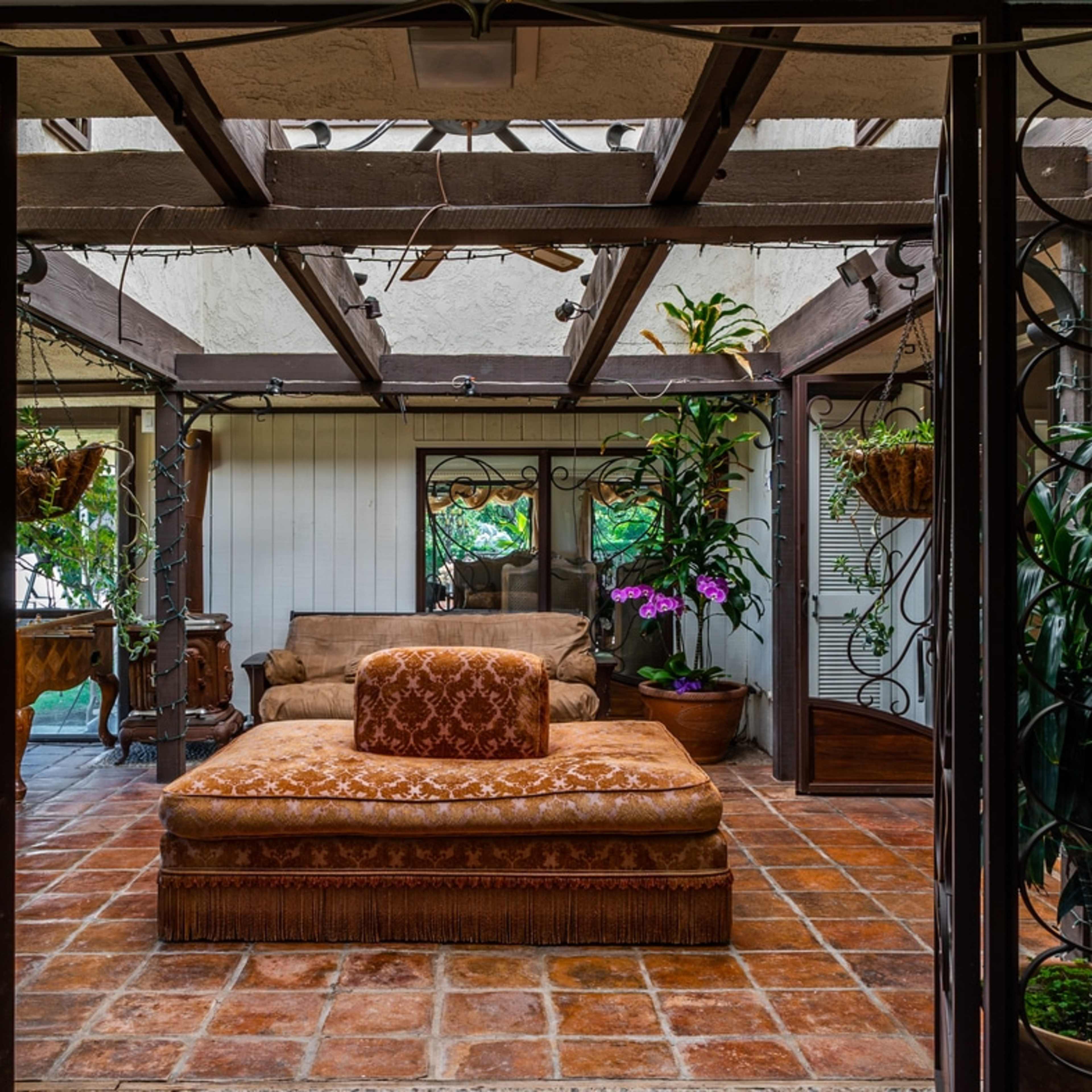 A sunlit patio surrounded by plants, featuring a large, patterned sofa on a tiled floor.