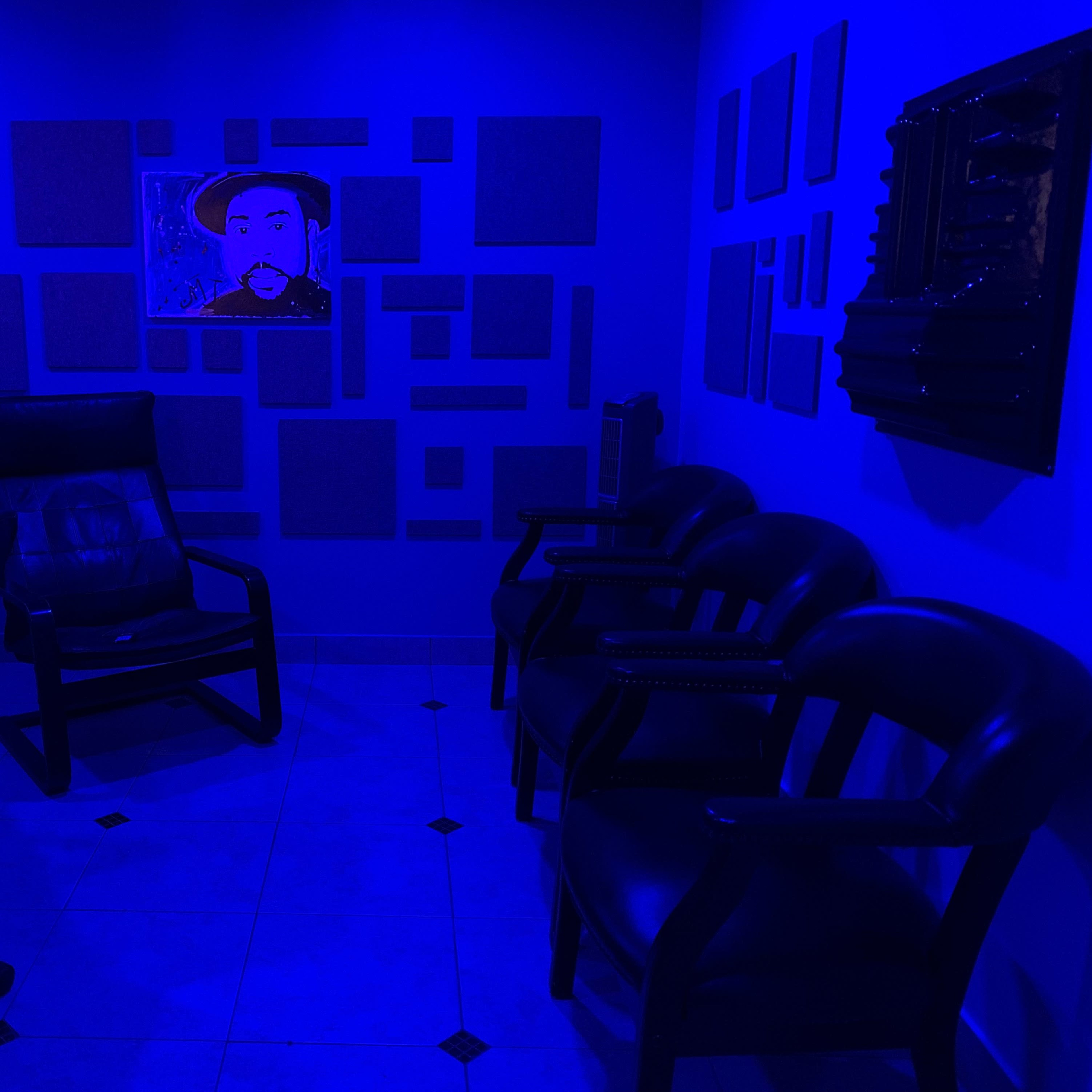 A dimly lit room with blue walls featuring soundproof panels, a painting on the wall, and several black chairs arranged around a table.