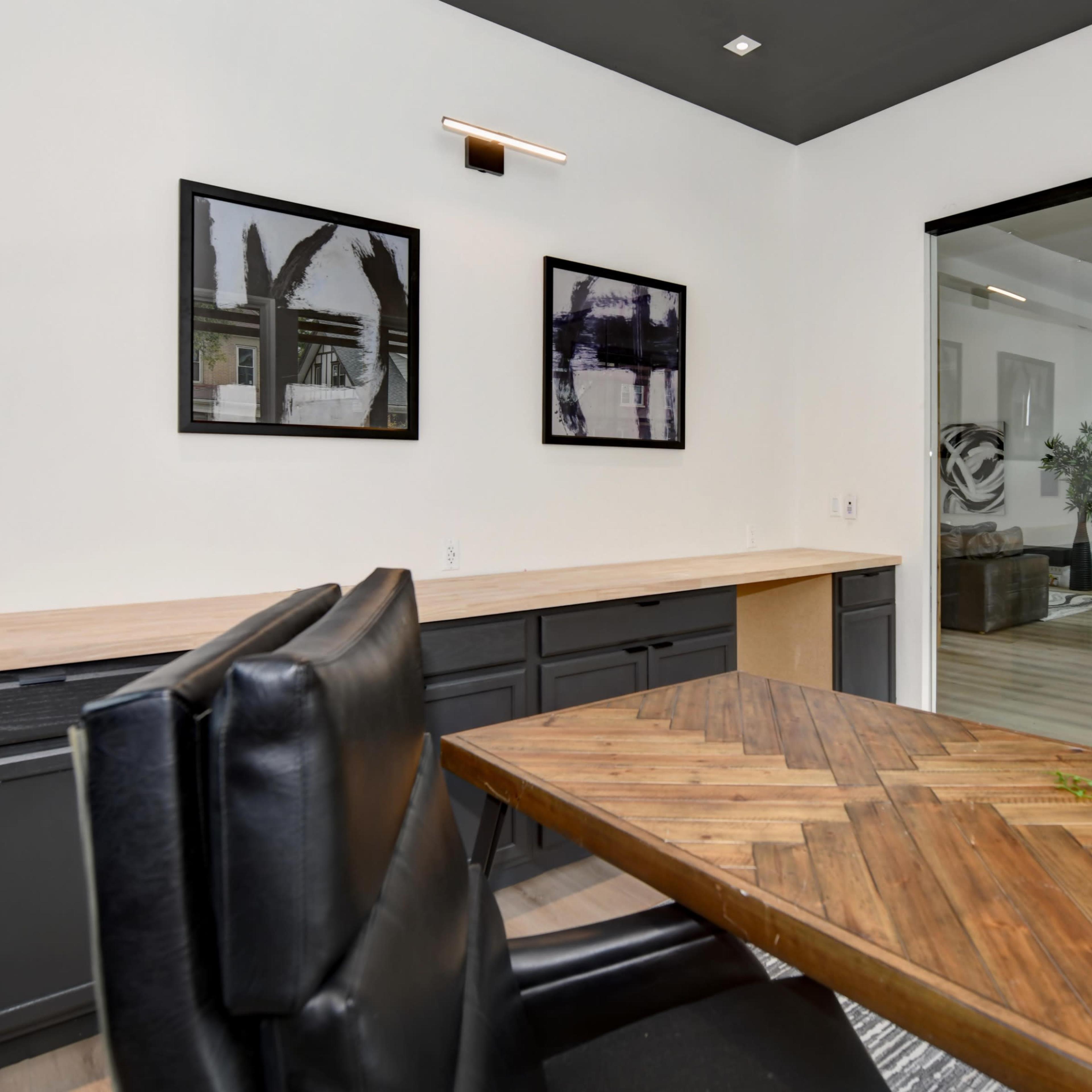 A modern interior features a wooden table with a chevron pattern, black leather chairs, and framed wall art, alongside a glass-walled staircase.