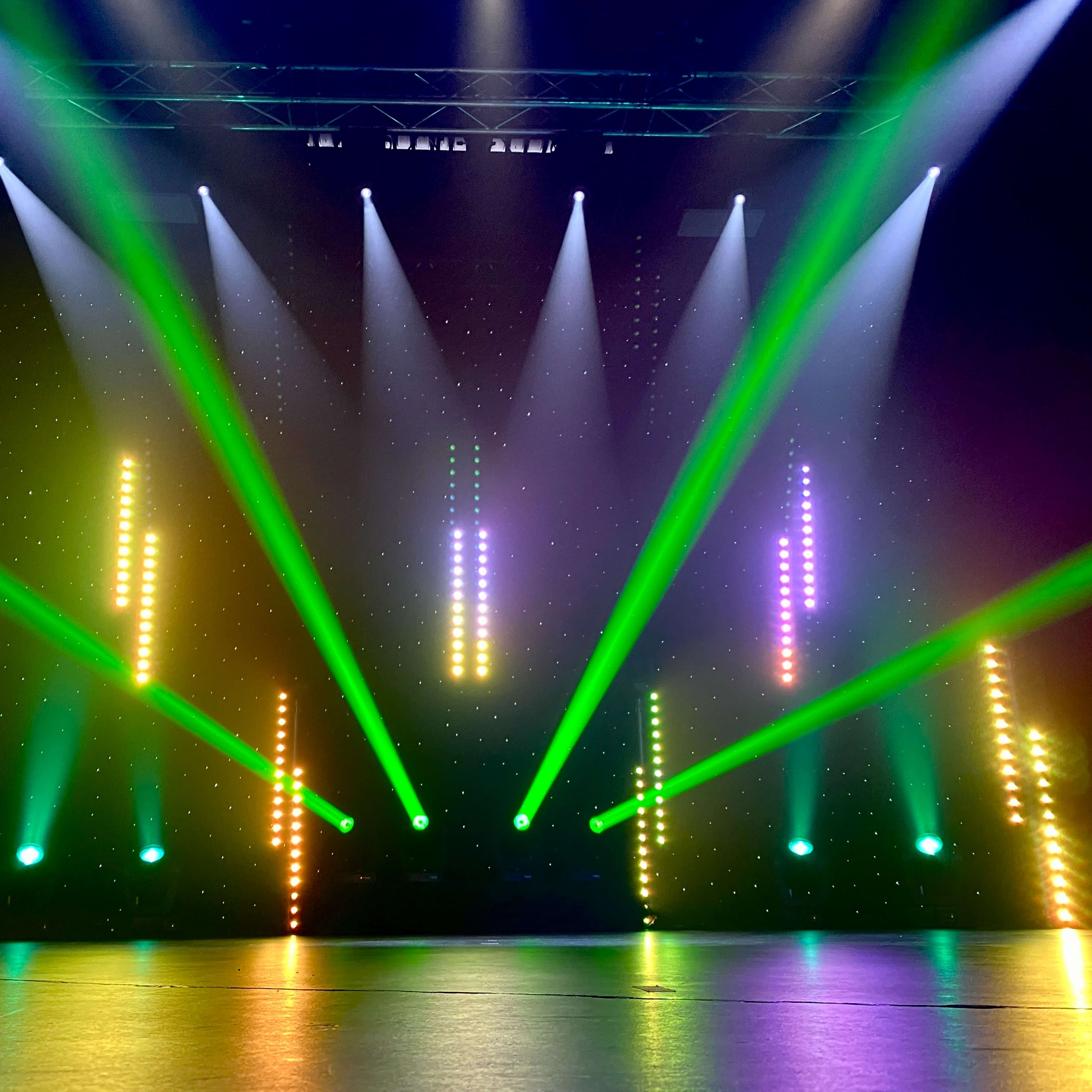 A stage illuminated by vertical light beams in green, yellow, and purple, with a dark backdrop and a shiny floor.