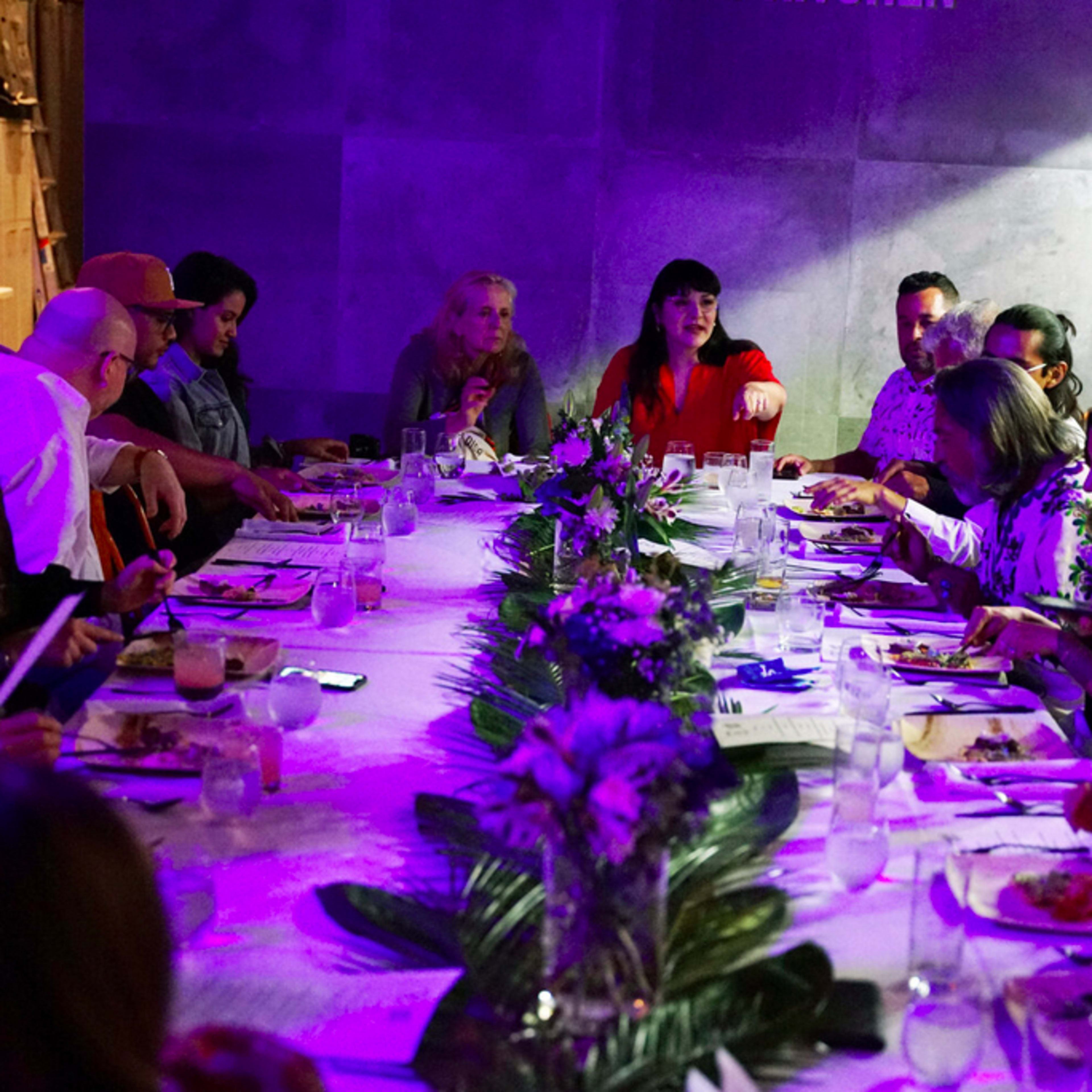 A large group of people is seated around a long table, engaging in conversation while dining in a dimly lit room with purple lighting.