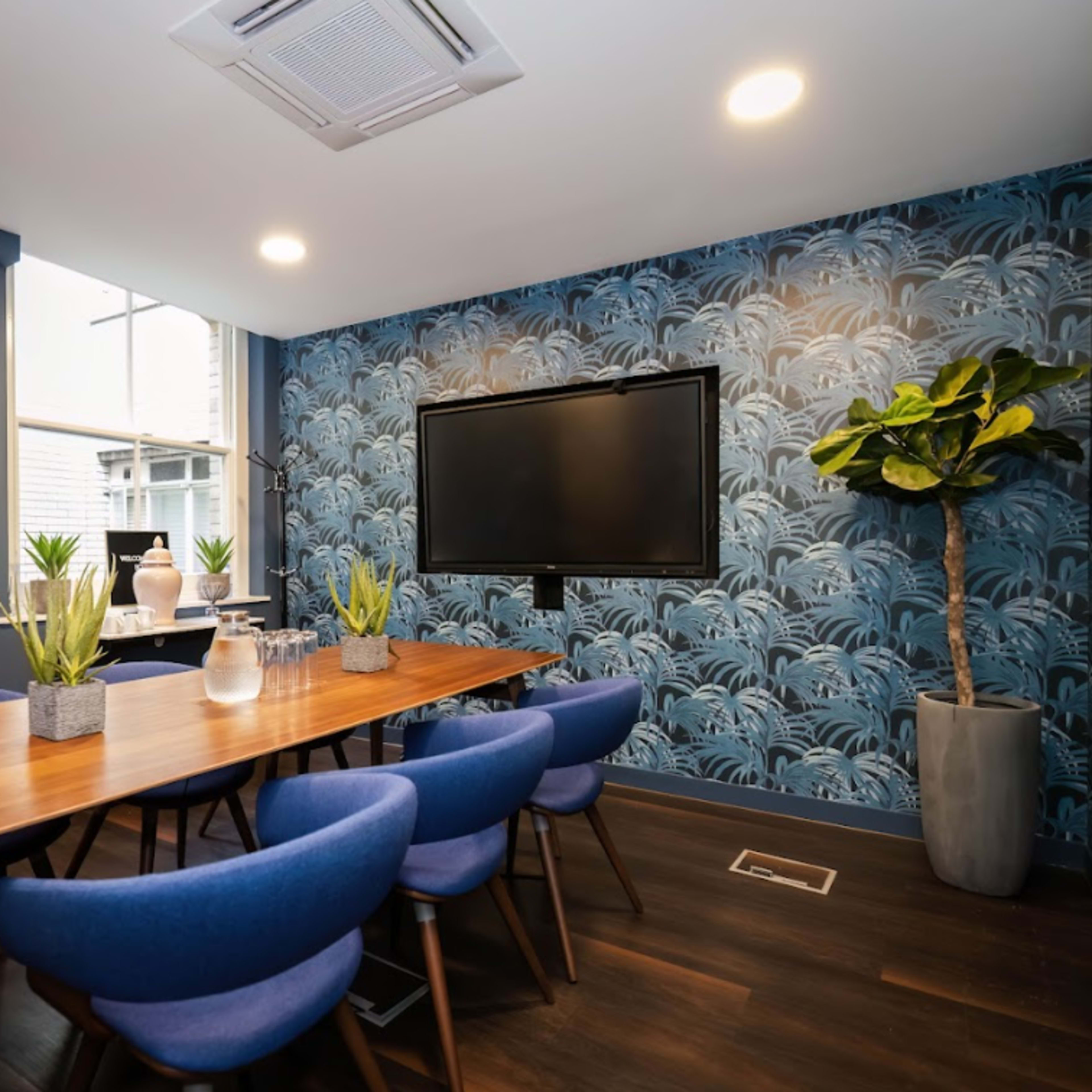 A modern conference room featuring a wooden table surrounded by blue chairs, a wall with botanical wallpaper, a television mounted on the wall, and decorative plants.
