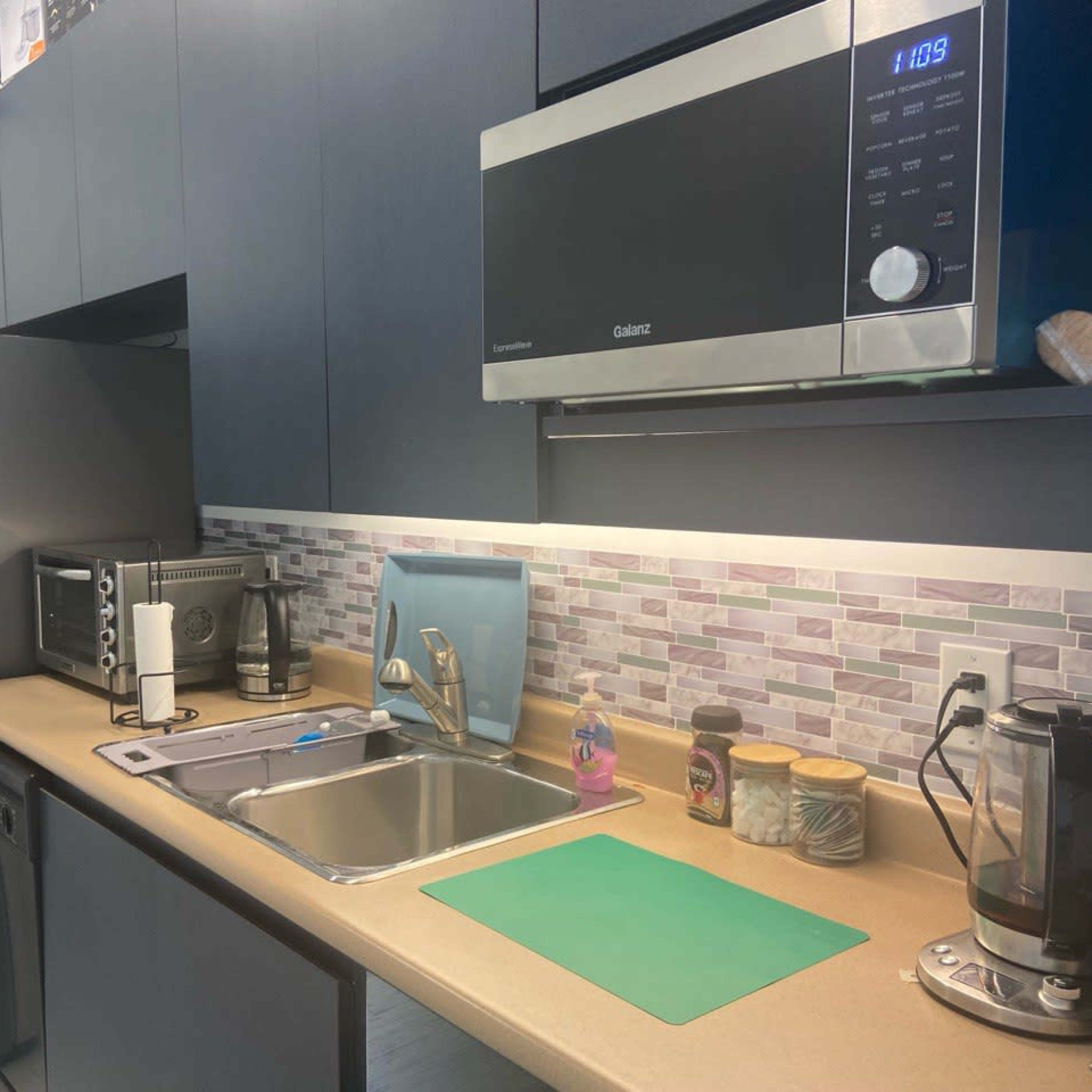 A modern kitchen with dark cabinetry, a stainless steel refrigerator, a microwave, a sink, and various countertop appliances.