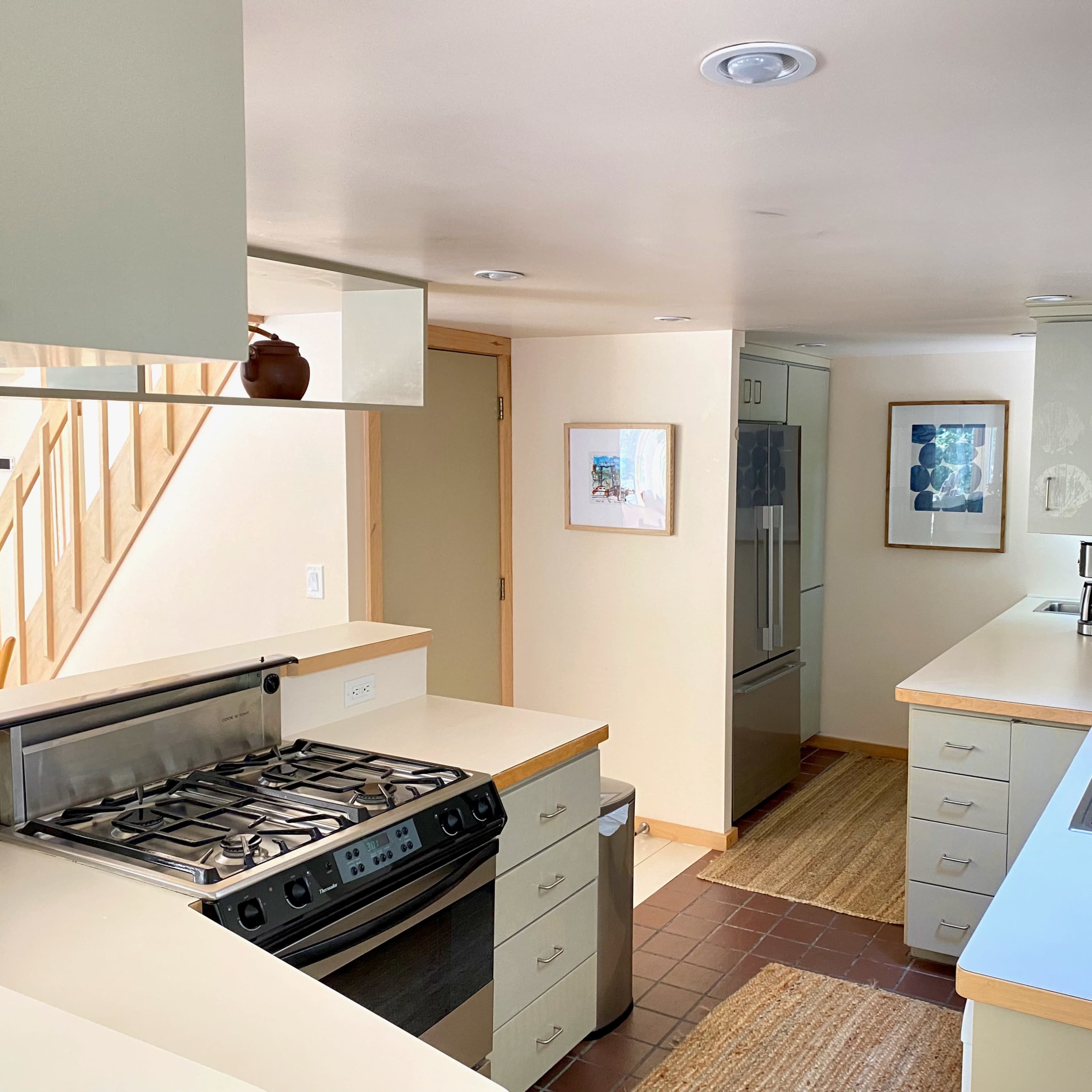 A modern kitchen with a gas stove, sink, and large refrigerator, featuring light-colored cabinets and a staircase in the background.