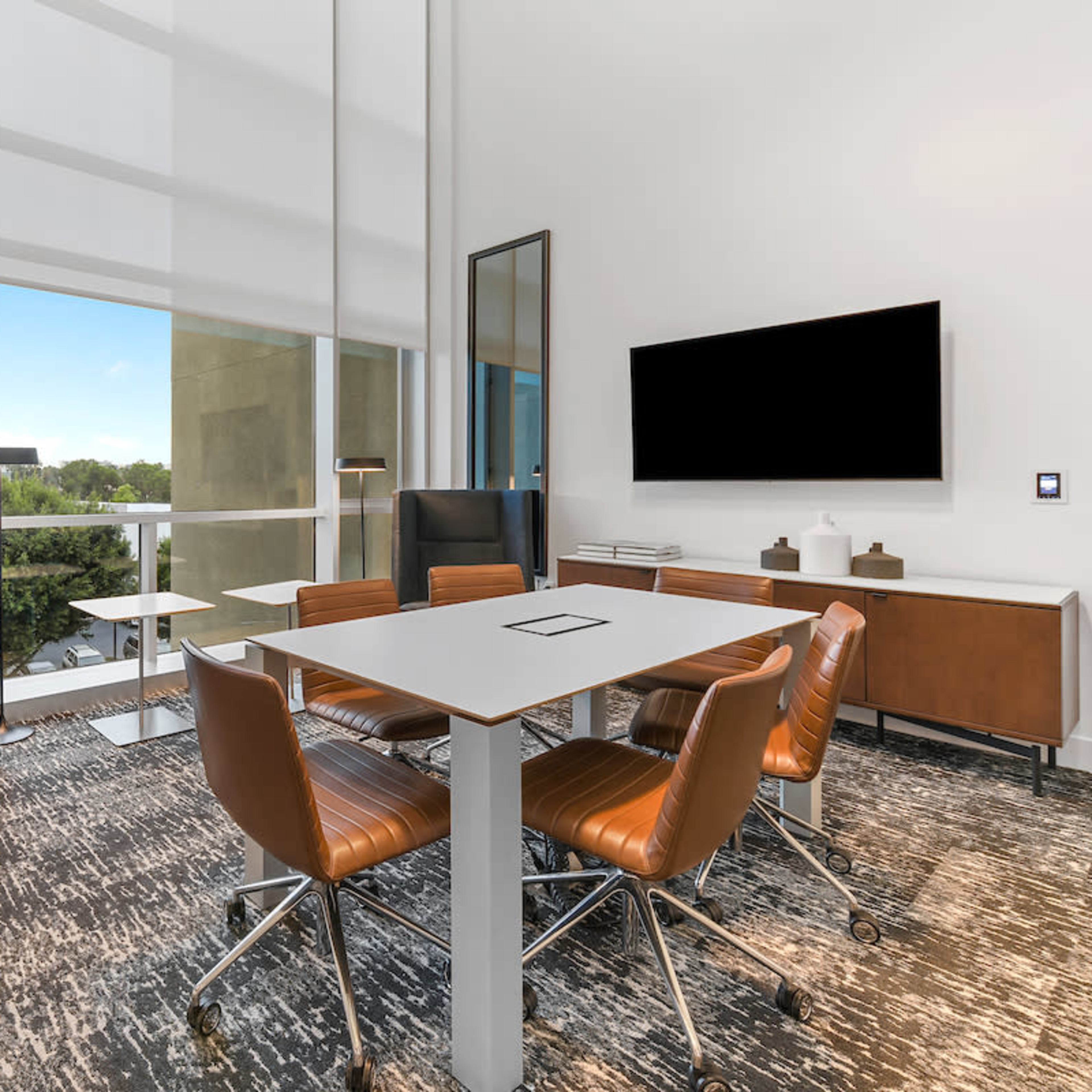A modern meeting room with a large window, featuring a table surrounded by chairs, a lounge area with a high-backed chair, and a wall-mounted television.