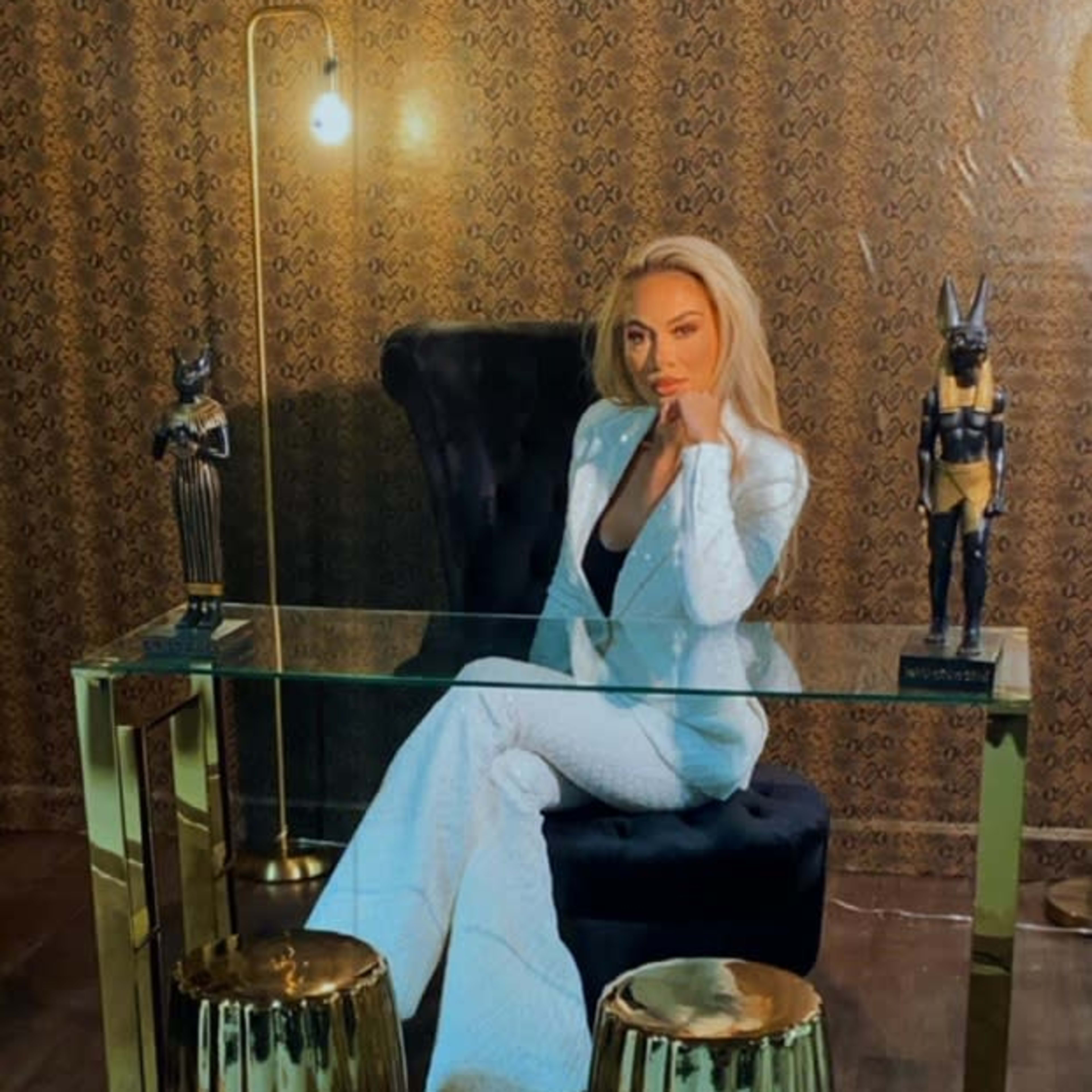 A woman in a white suit sits at a glass desk in front of a decorative wall, flanked by two golden stools and statues.