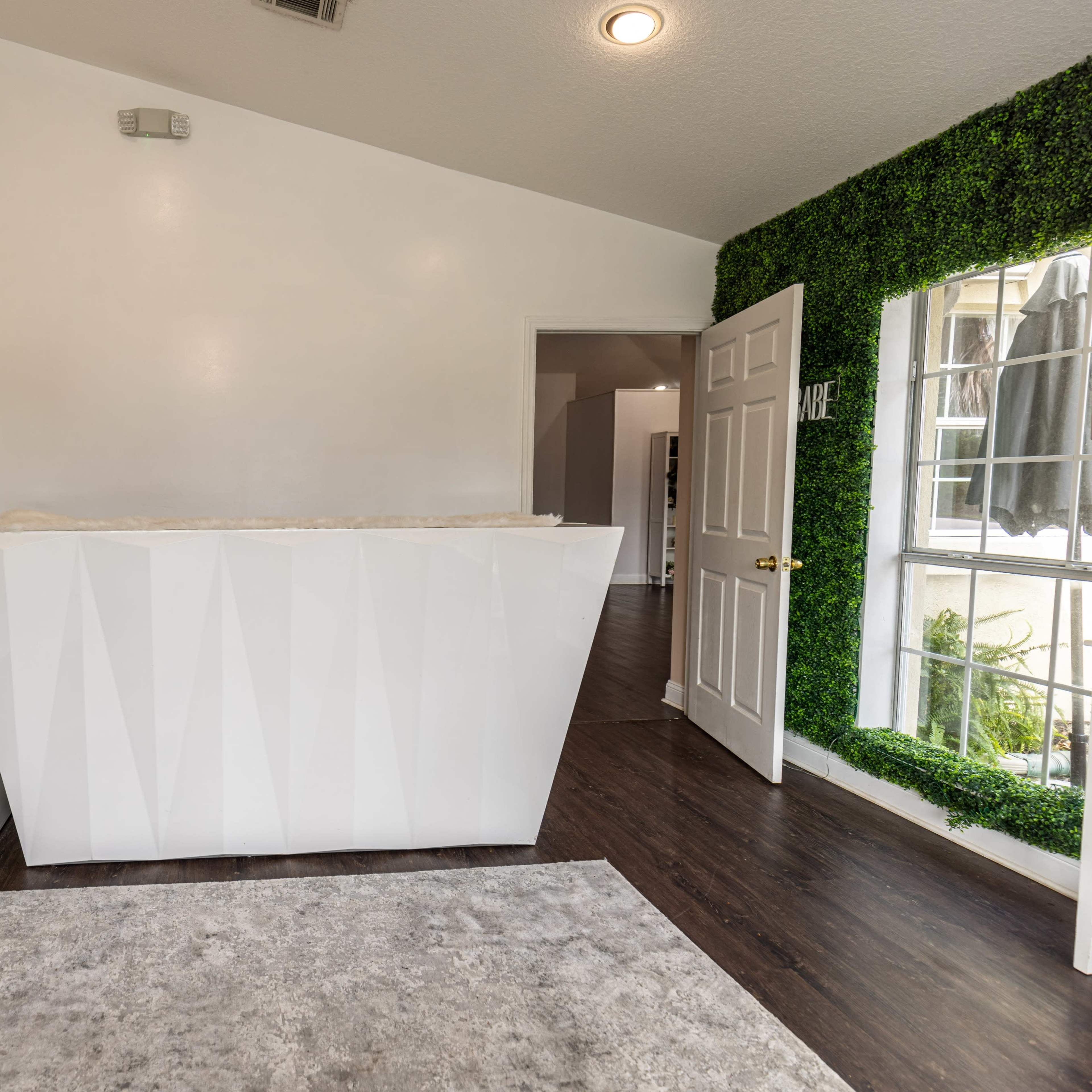 A modern reception area featuring a white geometric desk, a shelving unit, and a wall adorned with green foliage, with a door leading to another room in the background.