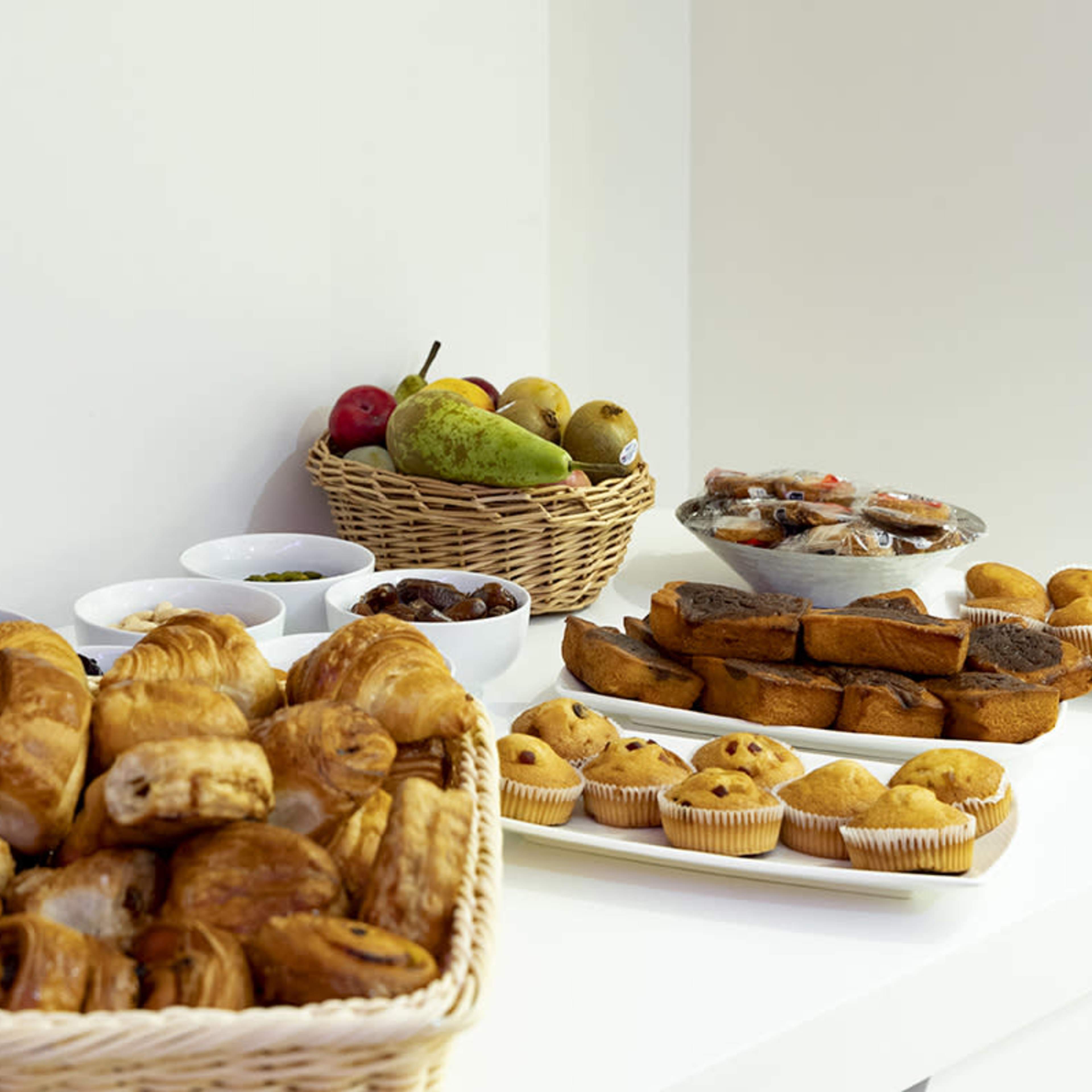 A variety of baked goods and fresh fruits are arranged on a white table, including croissants, muffins, and a selection of fruits in baskets.