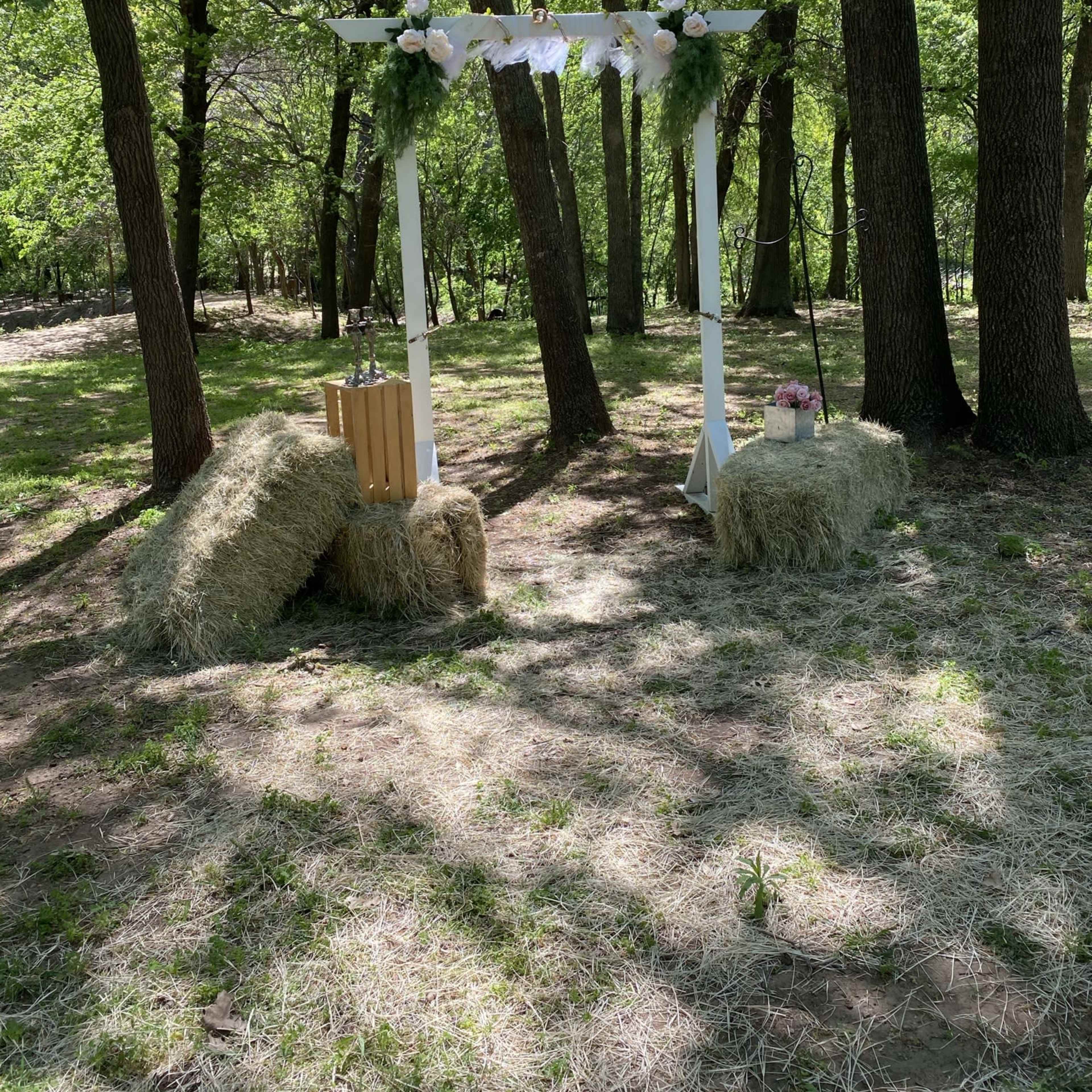 A white wedding arch decorated with flowers stands between two bales of hay in a wooded area.