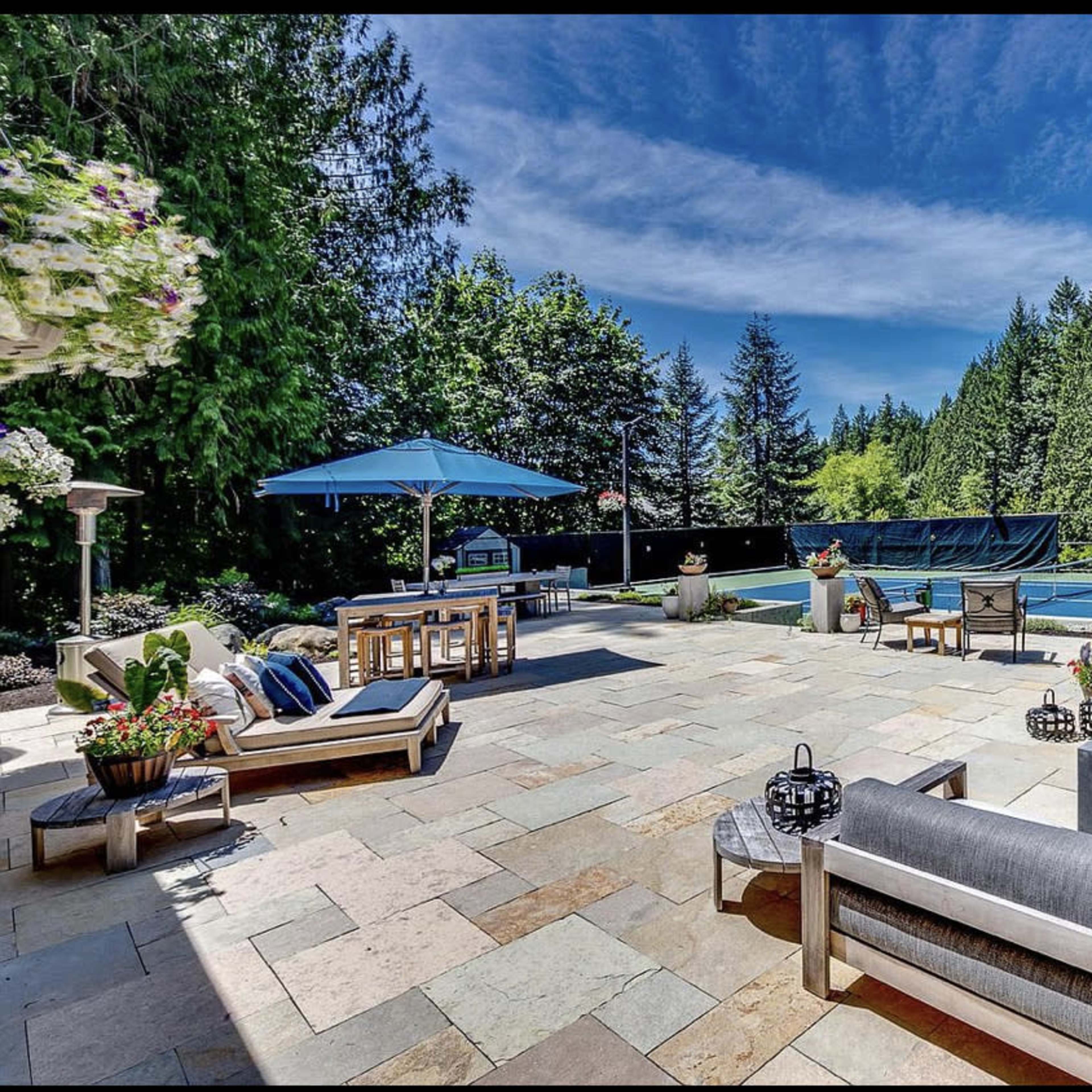 A spacious outdoor patio area with seating, a fire pit, and a tennis court surrounded by trees.