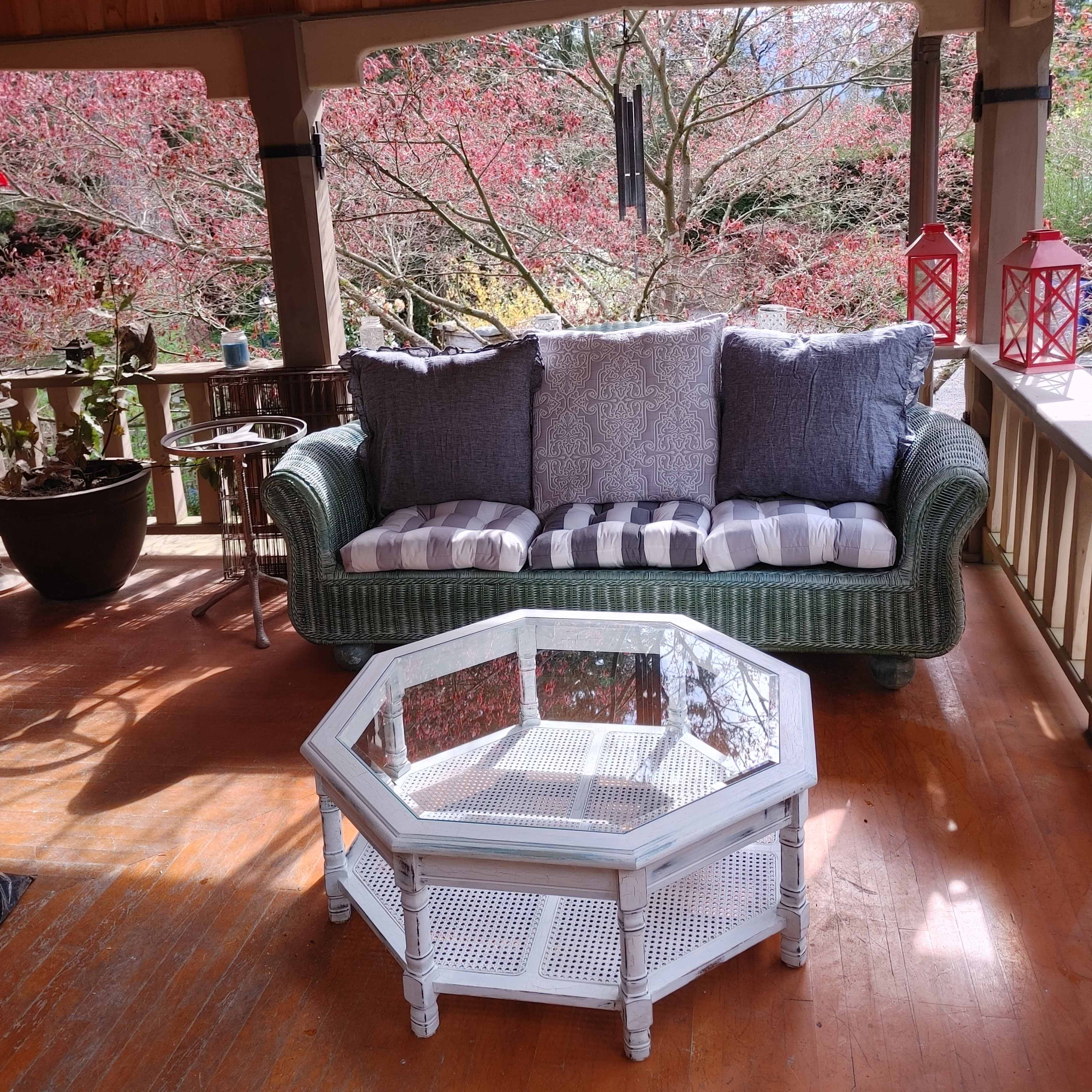 A wicker sofa with cushions is positioned beside an octagonal white coffee table on a porch, overlooking trees with pink foliage.