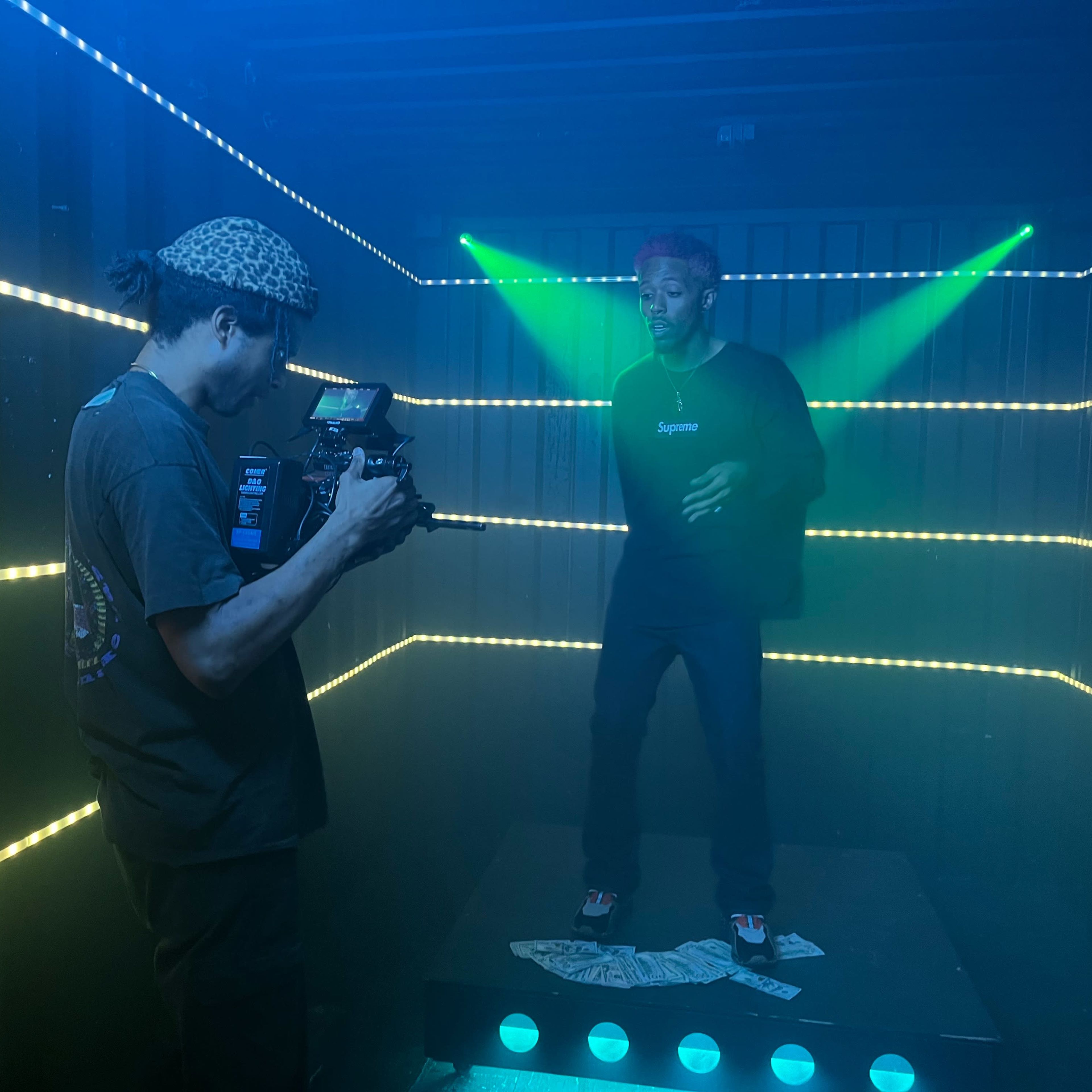 A person operates a camera while another stands on a platform surrounded by colorful lights and fog.