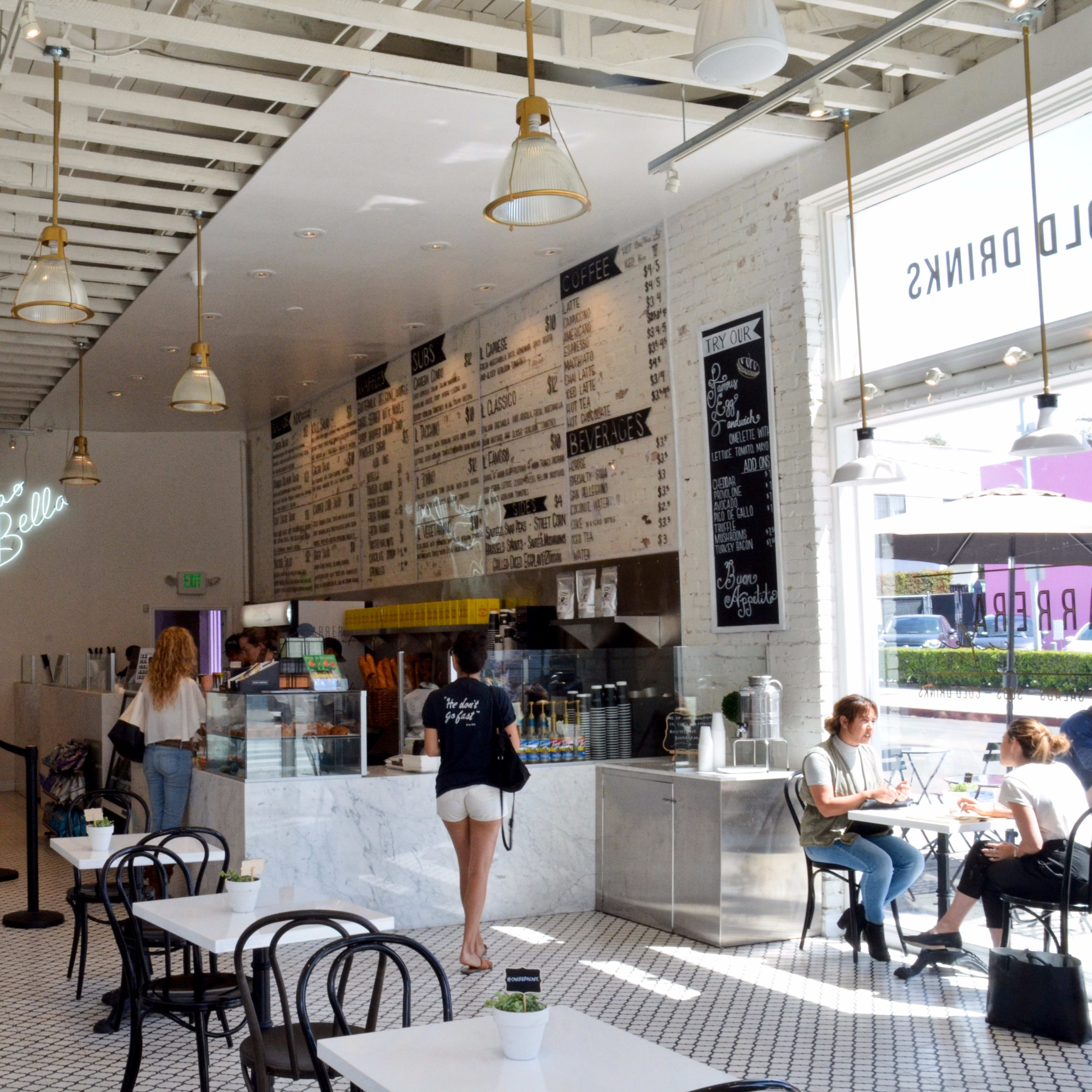 A bright, modern café interior with white walls, black and white tiled flooring, and patrons ordering at a marble counter.